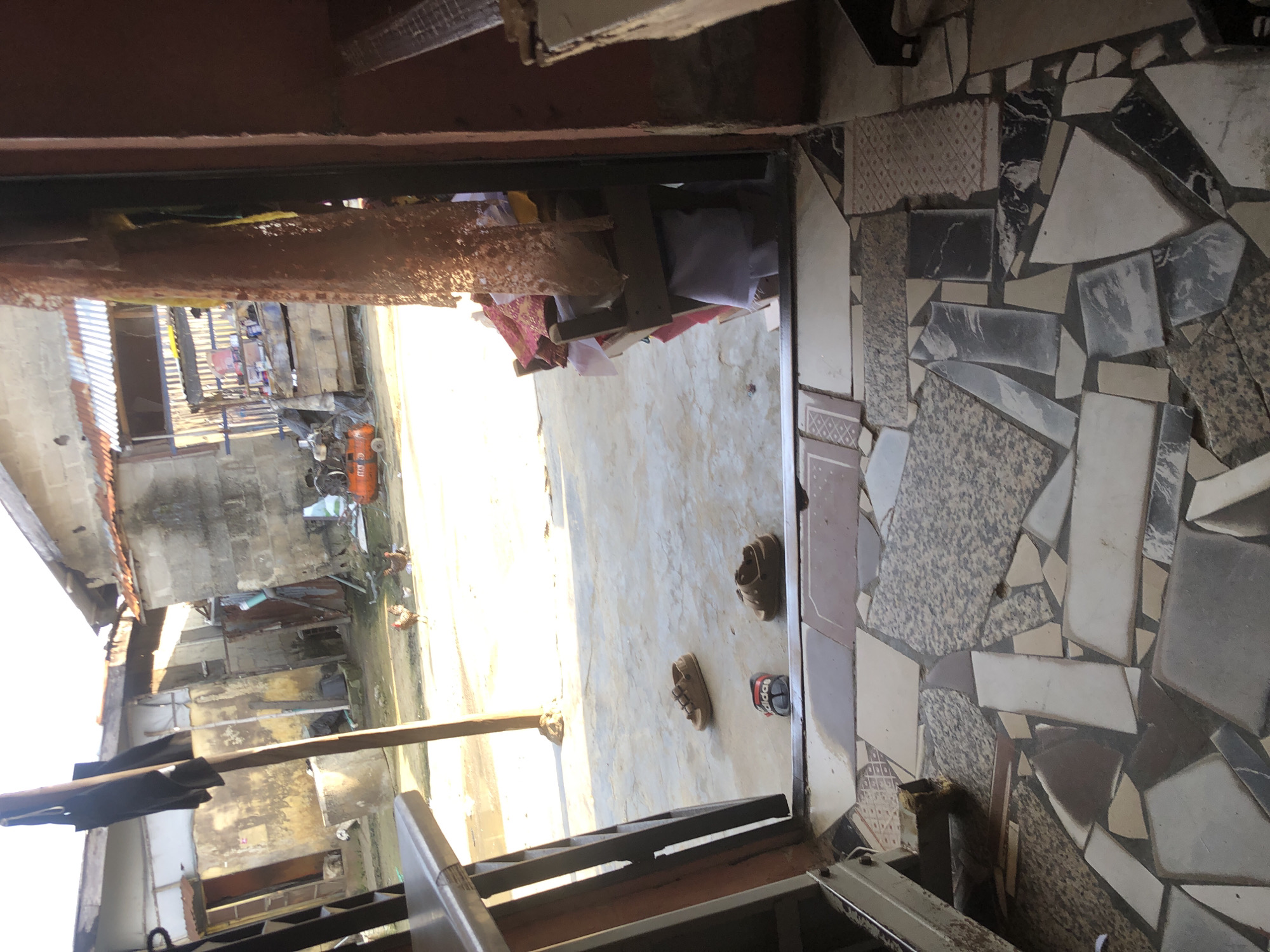 The image depicts an outdoor courtyard area, likely in Ado, Nigeria, with a focus on the immediate foreground and a glimpse into the background. The foreground consists of a mosaic tiled floor, with irregular shapes and varying patterns of stone and ceramic tiles. A large, roughly textured, reddish-brown pillar is prominently featured on the right side, partially obscuring the view into the background.
In the middle ground, several pairs of sandals are scattered on a concrete surface. A small, dark-colored cat or small animal is visible in the lower left corner of the frame, partially obscured by shadows. Beyond this, the courtyard opens up to reveal a backdrop of modest buildings constructed from concrete blocks and corrugated metal roofing. In the background, several chickens are seen roaming freely on the ground. An orange air compressor is also visible amongst some assorted items. The scene is illuminated by natural sunlight, creating distinct shadows. The overall impression is one of a functional, lived-in outdoor space.