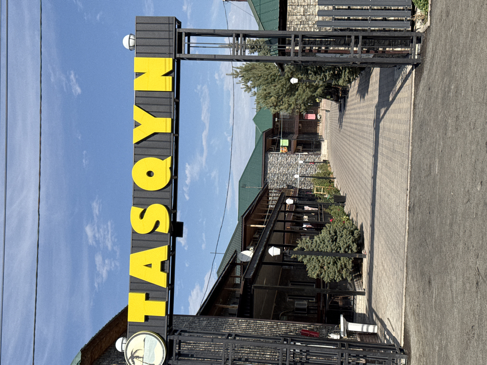 The image shows the entrance to a complex, likely a resort or hotel, identified by a large yellow sign overhead that reads "TASQYN". The sign is positioned above a black metal archway. To the left of the sign, a circular emblem featuring a palm tree and a beach scene is visible.
The archway leads to a paved walkway lined with trees and bushes. On either side of the walkway, there are buildings constructed with stone and wood, featuring balconies and green roofs. The buildings appear to be multi-story. Several lights, designed to resemble traditional lampposts, are placed along the walkway. Laundry is seen hanging from the balconies of the buildings.
The scene is set outdoors under a bright, partly cloudy blue sky. The ground in front of the archway is asphalt, transitioning to patterned paving stones further inside the complex. The overall impression is of a well-maintained establishment. The context provided indicates this is the location in Zharbulak, Kazakhstan. There are no people visible in the image.