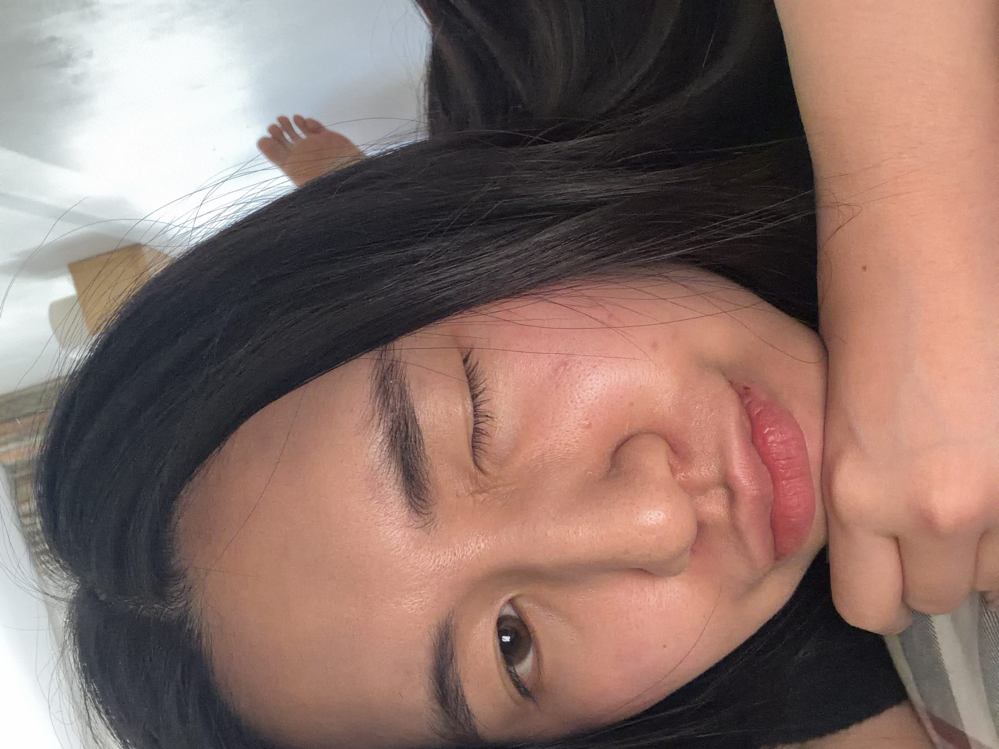 A close-up, tilted, color photograph shows a woman's face pressed against her arm. Her dark hair is spread around her head. She has one eye open and one closed. Her lips are slightly pursed. Part of a foot is visible in the upper left corner of the frame, and the background is nondescript. The location cannot be determined from the image.