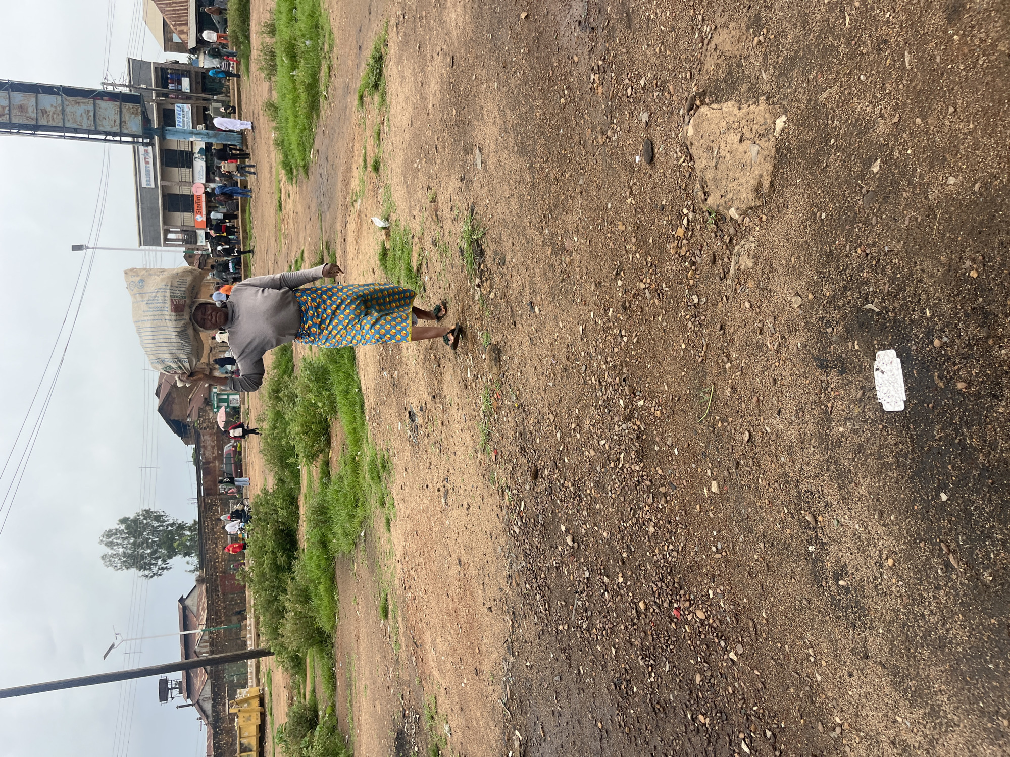 A woman is walking on a dirt road in Bukuru, Nigeria, carrying a large bag of goods on her head. The road is unpaved and appears to be in a commercial or residential area. In the background, there are several buildings, including what appears to be a market or shops. There are also other people in the background, some of whom are walking or standing around. The sky is overcast, suggesting it may be cloudy or raining. The woman is wearing a grey long-sleeved shirt and a colorful patterned skirt. She is also wearing sandals. The bag on her head is large and appears to be made of woven material. The scene depicts everyday life in a Nigerian city.