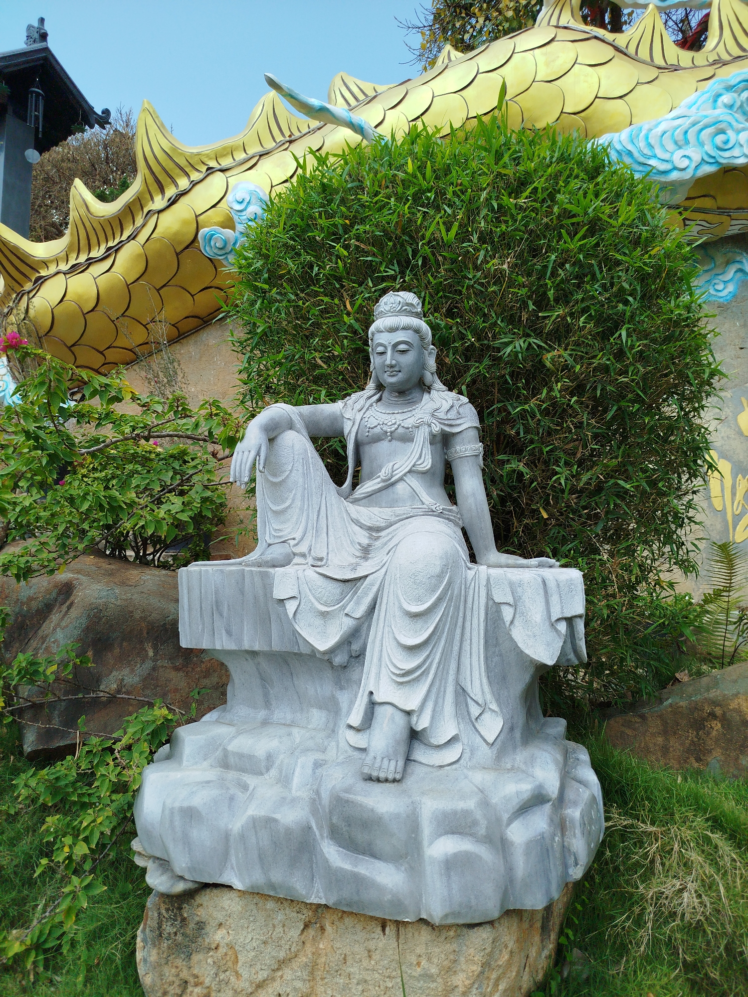 The central focus of the image is a serene gray stone statue of a deity, likely Guanyin (Kuan Yin), seated in a relaxed posture. The statue features intricate carvings, including a detailed crown, necklaces, and draped robes. One leg is bent with an arm resting on the knee, while the other leg extends downwards. The deity's face conveys a peaceful and calm expression.
Behind the statue, a large, golden dragon sculpture with prominent scales and spikes forms a striking backdrop. Sections of the dragon are adorned with light blue, cloud-like patterns. Lush green foliage, including bamboo and other bushes, surrounds the statue and extends into the mid-ground. Small clusters of pink flowers are also visible among the greenery on the left side. To the far left, a portion of a dark-roofed building, possibly a temple structure with wind chimes hanging from its eaves, is visible against a clear, light blue sky. The ground around the statue consists of natural rocks and green grass.
The scene depicts a tranquil outdoor setting, characteristic of a temple garden or religious site, located in Diom A, Vietnam. The lighting indicates a bright daytime, likely morning or early afternoon, under a clear sky. There is no discernible activity taking place, and no readable text is visible within the image.