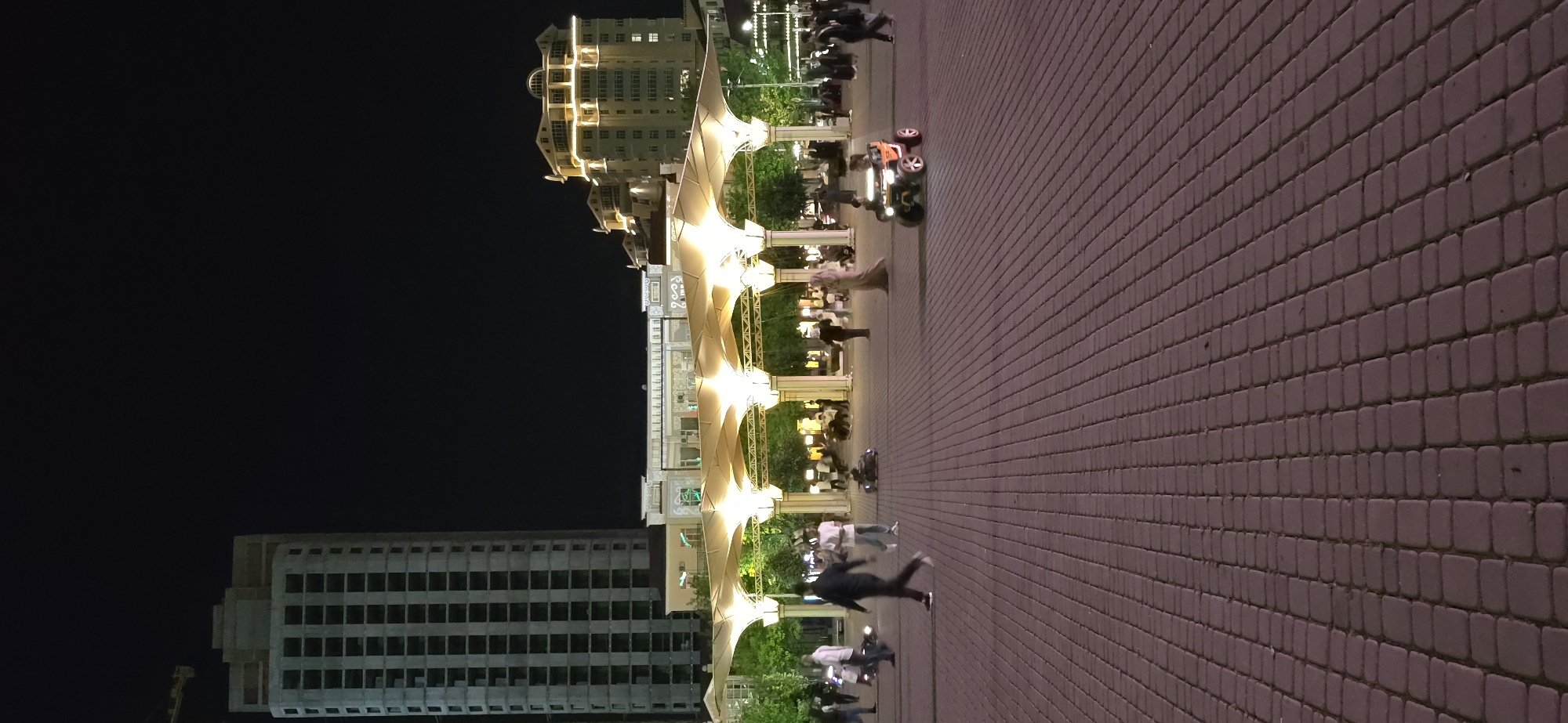 The image shows a nighttime scene in Grozny, Russia. The foreground features a wide, brick-paved pedestrian area. A series of illuminated, arched structures runs along the left side, with trees and people visible within and around them. Several people appear blurred, indicating motion. In the background, there are buildings of varying heights, one under construction and another with illuminated windows. The sky is dark.