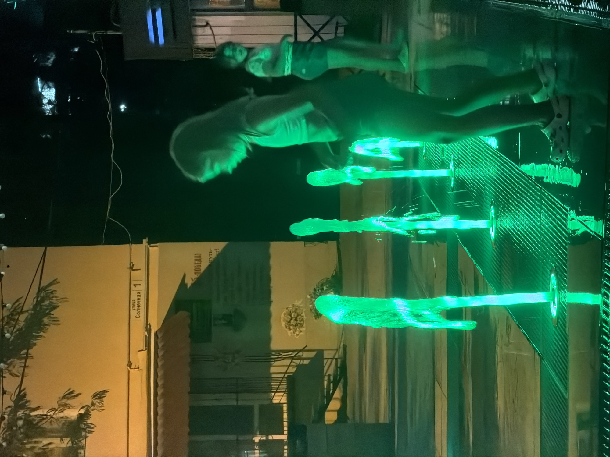 In the city of Partenit, Ukraine, two young girls are captured in motion near a contemporary fountain. The fountain features multiple illuminated water jets, casting a vibrant green glow on the surrounding wet pavement and the girls. The girl in the foreground, with long dark hair, is silhouetted against the lights, wearing a light-colored top and dark shorts. She appears to be walking or dancing, with her legs in motion. Behind her, a second girl, also with dark hair, is partially visible, wearing a patterned top and shorts. The background reveals a building with a yellow facade and a red awning, some of which is illuminated by artificial light. A street sign in the upper left reads "ул. Солнечная 1" (Sunny Street 1). The overall scene is nocturnal, with the green light of the fountain dominating the atmosphere.