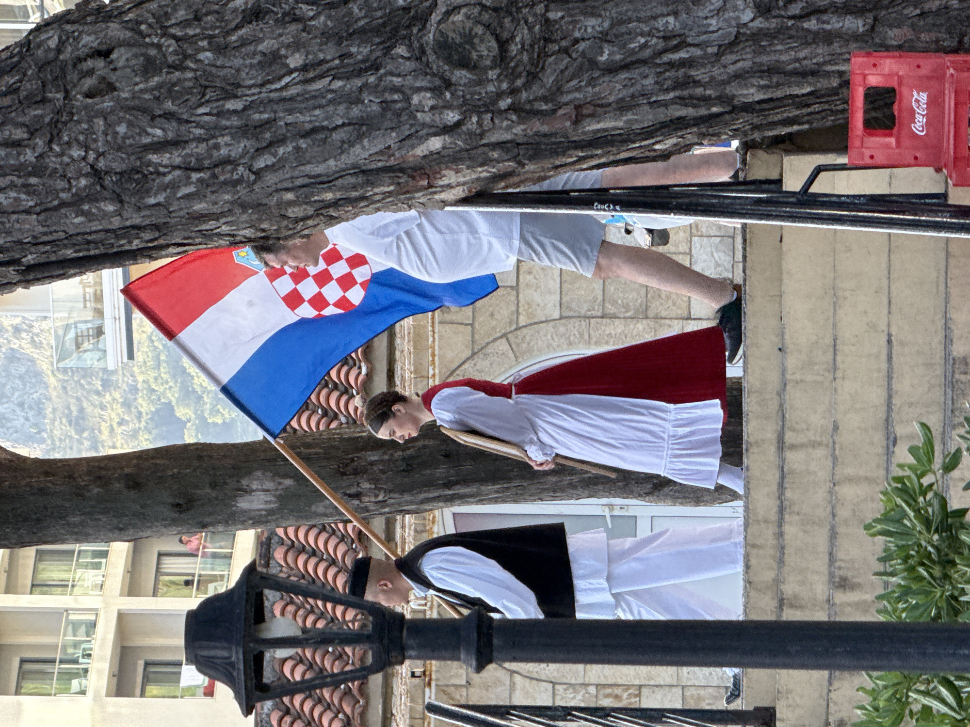 The image depicts an outdoor scene in Budva, Montenegro, featuring multiple individuals and architectural elements. In the foreground, a prominent black, ornate street lamp post stands on the left. To its right, a large tree trunk with rough, textured bark occupies a significant portion of the right side, partially obscuring the view.
A group of people is ascending a stone staircase. On the far left, a man is dressed in traditional attire, consisting of a white long-sleeved shirt, a black sleeveless vest, and white trousers. He wears a dark, possibly black, cap and carries a wooden pole or staff over his right shoulder. Following him is a woman also in traditional dress, featuring a white long-sleeved blouse, a red skirt, and a white apron. Her hair is pulled back, and she holds a flat, paddle-like wooden object in her hands. Behind her, partially obscured by the tree trunk, a man in a white t-shirt and gray shorts walks, holding a Croatian flag (red, white, and blue with a checkerboard shield) aloft on a wooden pole. His left leg is visible, wearing a dark shoe. Below the steps on the far right, two red plastic Coca-Cola crates are stacked.
In the mid-ground, behind the individuals and the lamp post, red terracotta roof tiles are visible. Above these, a modern multi-story building with white facades and glass balconies is prominent. On one of the balconies on the third floor, a person in a pink shirt is visible leaning on the railing, observing the scene. Green foliage is present at the base of the staircase in the foreground. In the distant background, hazy green hills or mountains rise, characteristic of the Montenegrin landscape.