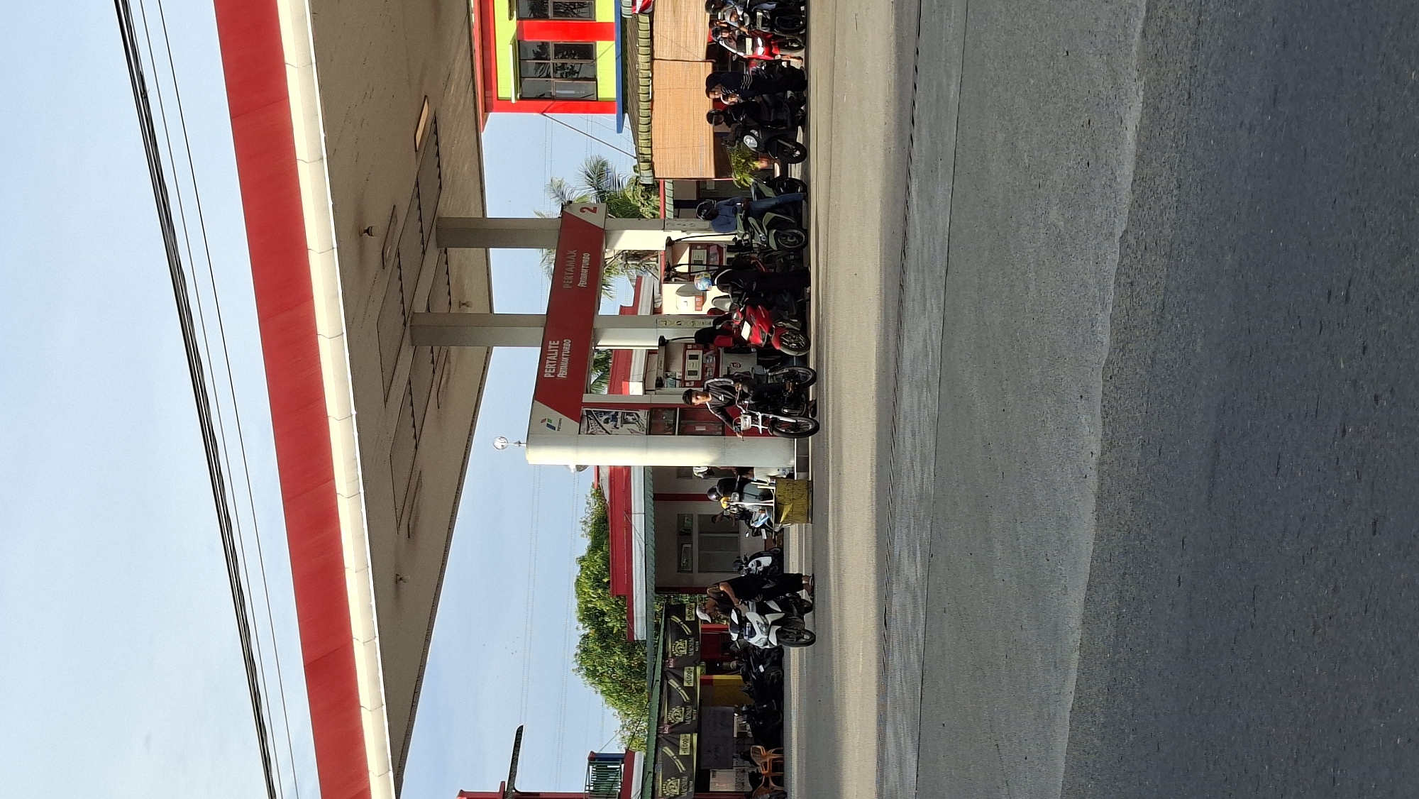 A busy gas station in Lhokseumawe, Indonesia, is depicted in this vertical, eye-level shot. The station features a red and white canopy extending over multiple gas pumps. Several people on motorcycles are waiting to refuel or have just refueled. In the foreground, a man on a white scooter is being attended to by another man. Behind them, a line of motorcycles and riders is visible. The background shows a store with red and green accents and various signs. The sky is clear, and the lighting suggests it's daytime. The overall impression is one of everyday activity at a local service station.