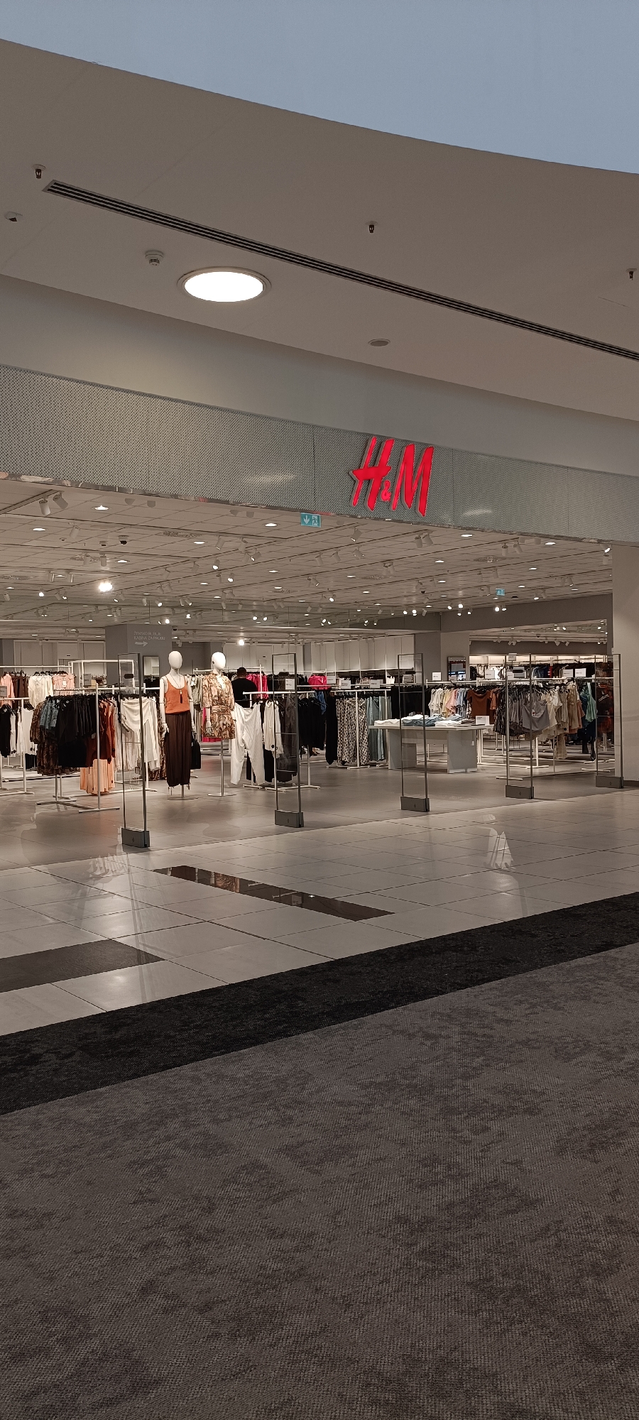 The image shows the exterior of an H&M clothing store, likely located in a shopping mall given the polished floor and indoor lighting. The H&M logo is prominently displayed in bright red neon letters above the store entrance. Inside the store, racks of clothing are visible, arranged neatly and illuminated by overhead lights. Mannequins are also present, displaying various outfits. The overall atmosphere appears to be calm and orderly, suggesting the store is either open for business or preparing to open. The time of day is unclear due to the indoor setting, but the lighting suggests it is either late evening or early morning, or the mall is artificially lit to mimic daylight. There are no visible signs of weather conditions. The floor in front of the store is tiled with a reflective surface, and a dark carpet runs along the bottom of the image, indicating a pathway or an area within the mall. No people are actively engaged in shopping or any other discernible activity. The scene depicts a commercial retail environment. Visible text includes the H&M logo and a directional arrow sign with text in a foreign language that is too small to read.