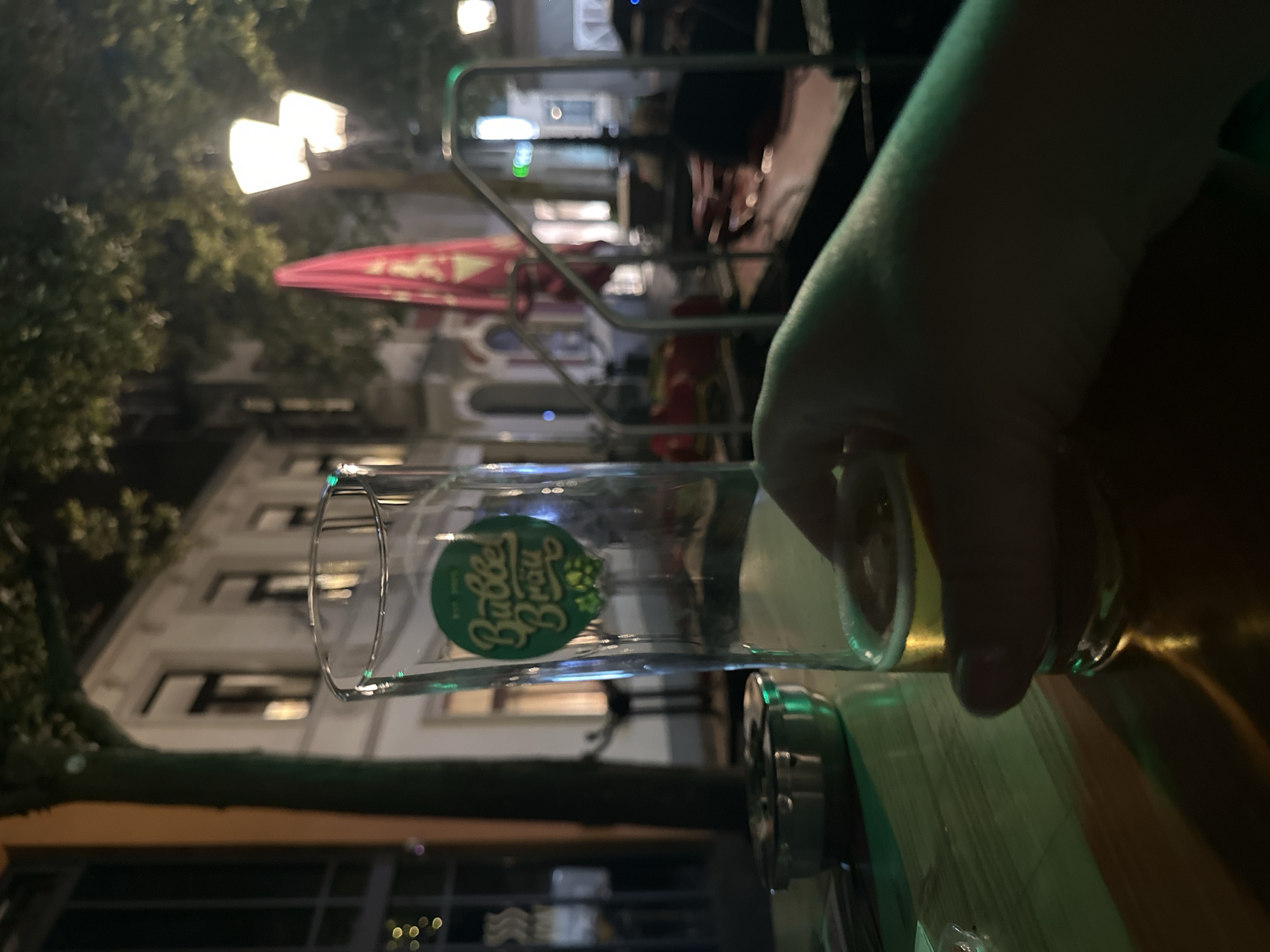 The image captures a close-up of a hand holding a glass of beer at night, presumably outdoors at a bar or restaurant. The glass has a green label with the text "Babbel Bräu" and images of hops. In the background, there are buildings with illuminated windows, trees, streetlights, and a red umbrella. The overall lighting suggests an evening setting. The environment appears to be a street in the city of Krefeld, Germany, with architectural elements consistent with German cityscapes. There are no other people clearly visible in the foreground or interacting with the subject. The focus is on the hand and the glass of beer.