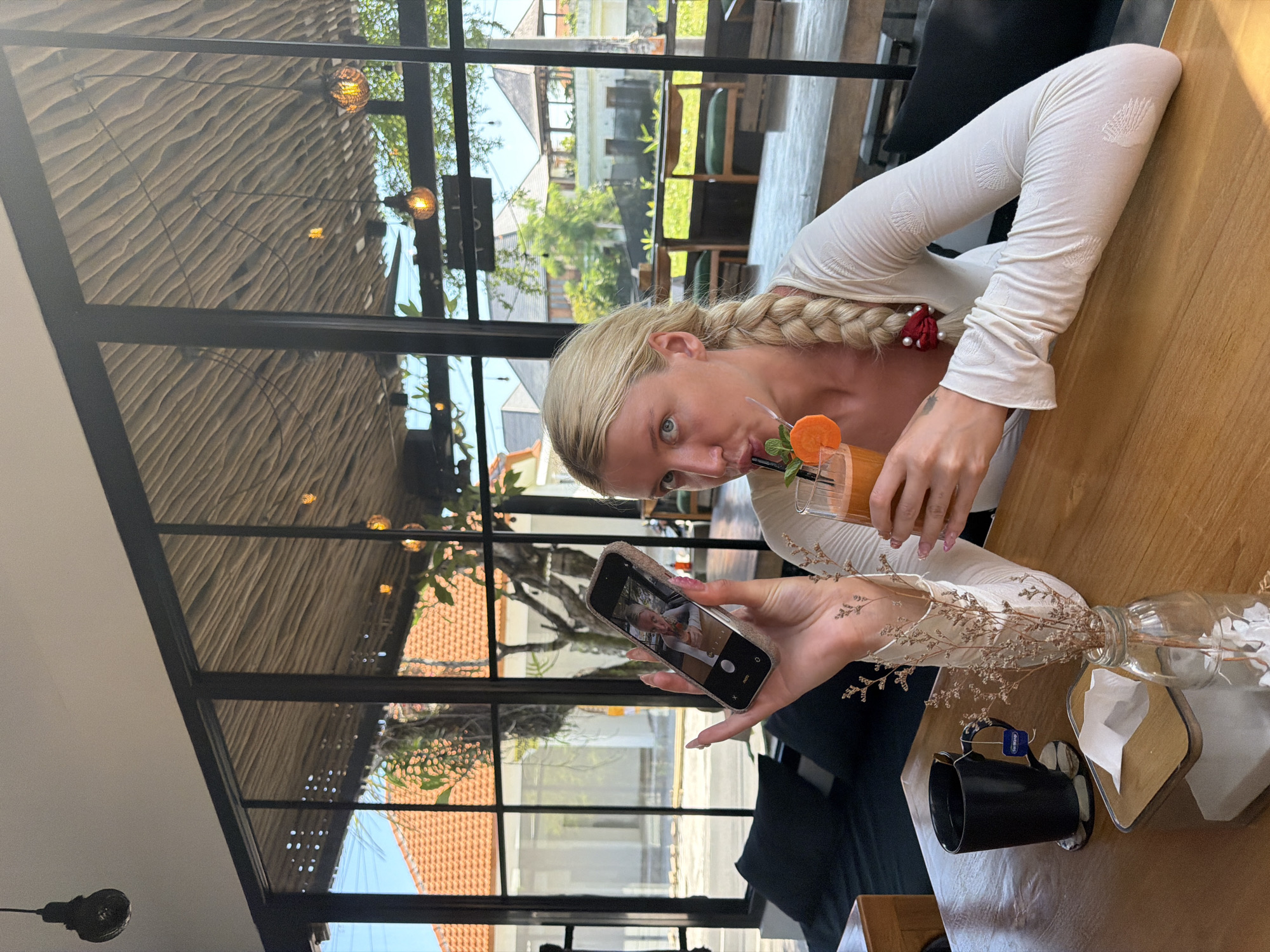 In North Kuta, Indonesia, a blonde woman with a braid is sitting at a wooden table. She is wearing a white long-sleeved shirt and holding a glass of orange juice with a slice of orange and mint. In her left hand, she is holding a smartphone, looking at the screen. The phone screen displays a picture of herself. On the table, there is a black mug with a tea bag, a tissue dispenser, and a small glass vase with dried flowers. The background shows a large window with a view of buildings with terracotta roofs and lush greenery. The ceiling above is made of woven material, and there are hanging light fixtures.