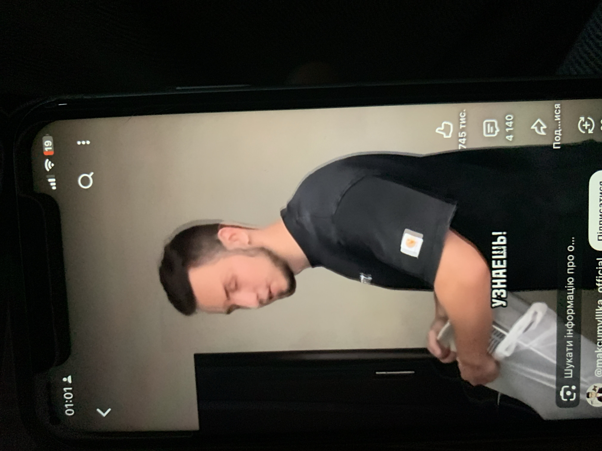 The image shows a man in a black t-shirt standing in front of a dark background. He is holding a clear plastic bag with white handles. The t-shirt has a small orange square logo on the sleeve. On the screen of a phone, which the image is of, there is text that says "УЗНАЕШЬ!". There are also icons indicating likes (745 тыс.), comments (4 140), and shares (Под...ися). The time displayed on the phone is 01:01. The location is not specified in the image, and there is no information to indicate it is in Zhmerynka, Ukraine.