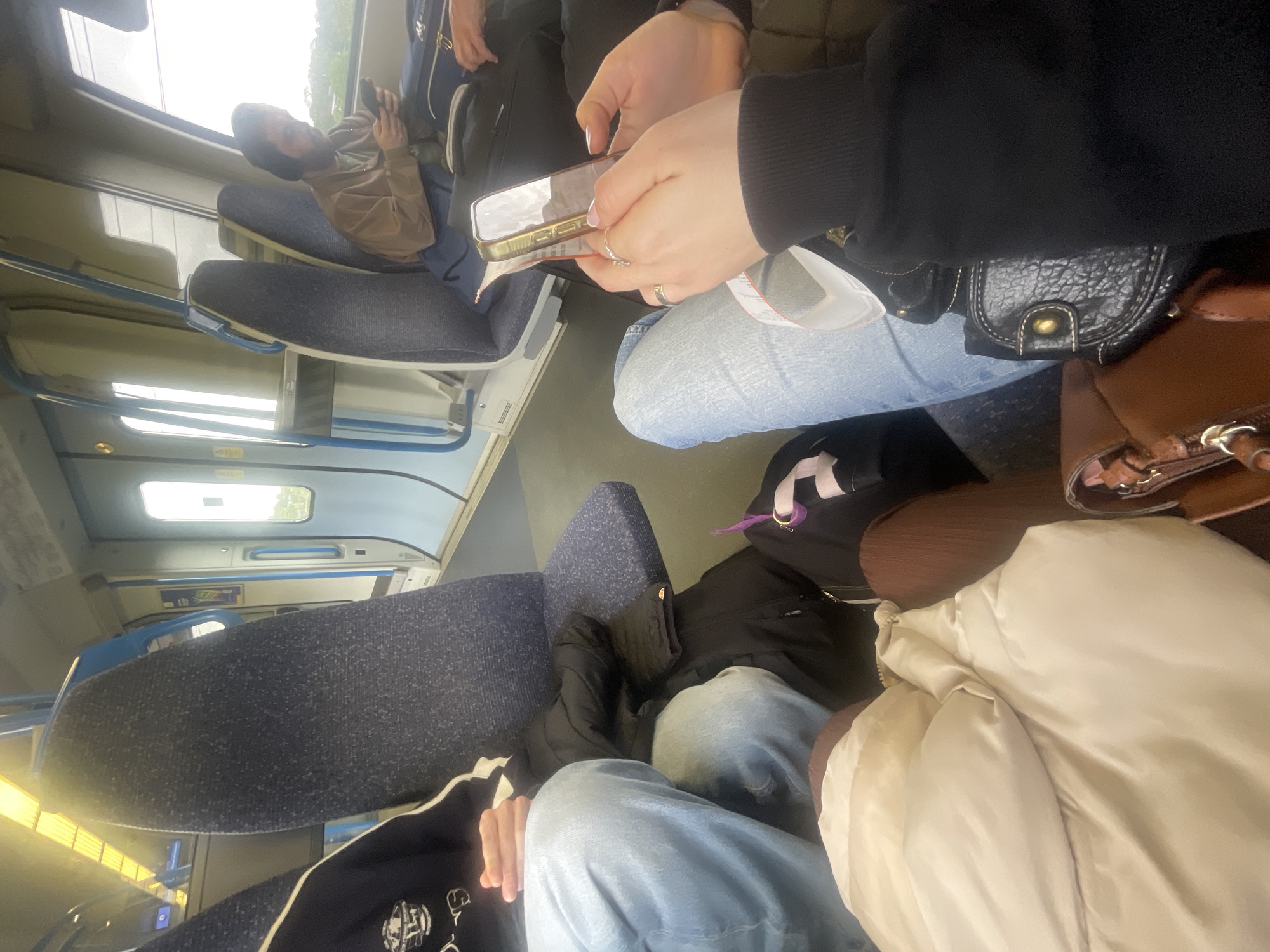 The image shows a first-person perspective of someone sitting on a train. The train's interior features blue upholstered seats with metal frames. Multiple passengers are visible, each occupied with their own activities. One person is looking at a phone, while the person taking the photo is holding a phone and a ticket. A black leather bag is beside the photographer. The setting is likely a commute, given the seating arrangement and the activity of looking at phones. The location is inferred as being on a train travelling in the city of St Albans, United Kingdom.