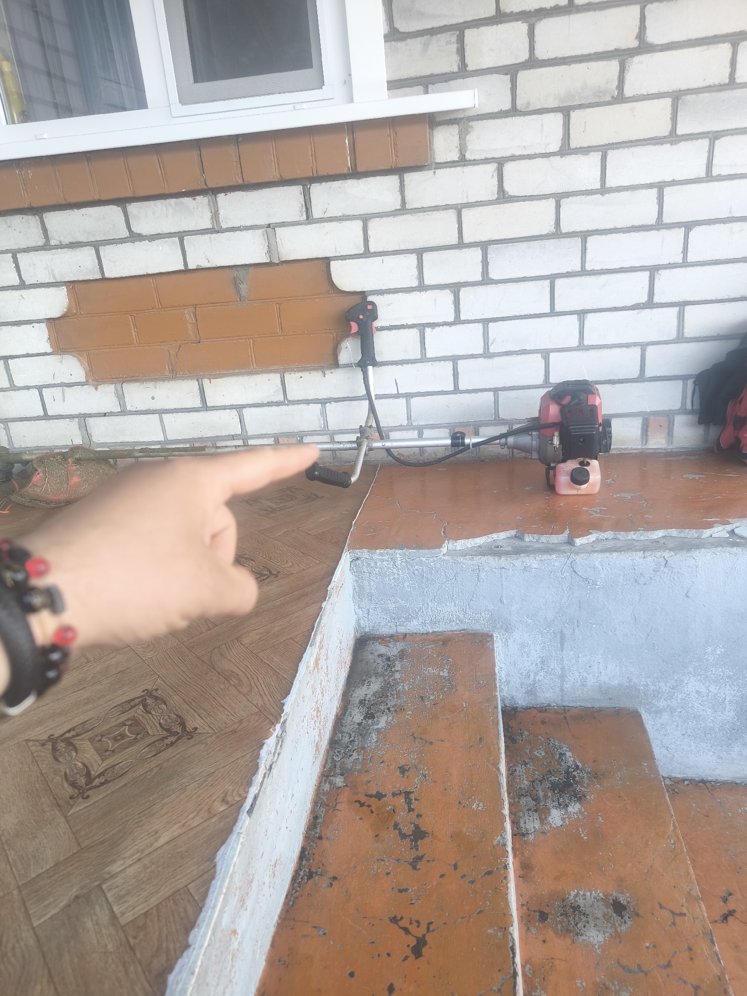 This image depicts an exterior scene at a structure in Unknown, Unknown, featuring a white-painted brick wall with sections of the original orange-brown brick visible. A white-framed window with a screen is present in the upper left. In the mid-ground, a gas-powered string trimmer (brush cutter) with a red and black engine, orange fuel tank, and silver shaft rests horizontally on an elevated, faded orange-brown concrete surface exhibiting significant wear and peeling paint. Grass clippings are visible near the trimmer's head on the left. To the lower left, a human left hand, wearing a dark beaded bracelet with red and black beads, points towards the trimmer. The flooring in the lower-left foreground is a wood-patterned linoleum with an ornate dark brown geometric design. To the lower right, a set of three weathered concrete steps, painted the same faded orange-brown with extensive chipping and dark discoloration, descends from the elevated surface. The vertical face of the steps is painted a light blue-grey. A dark fabric item is partially visible on the far right edge of the elevated concrete surface.