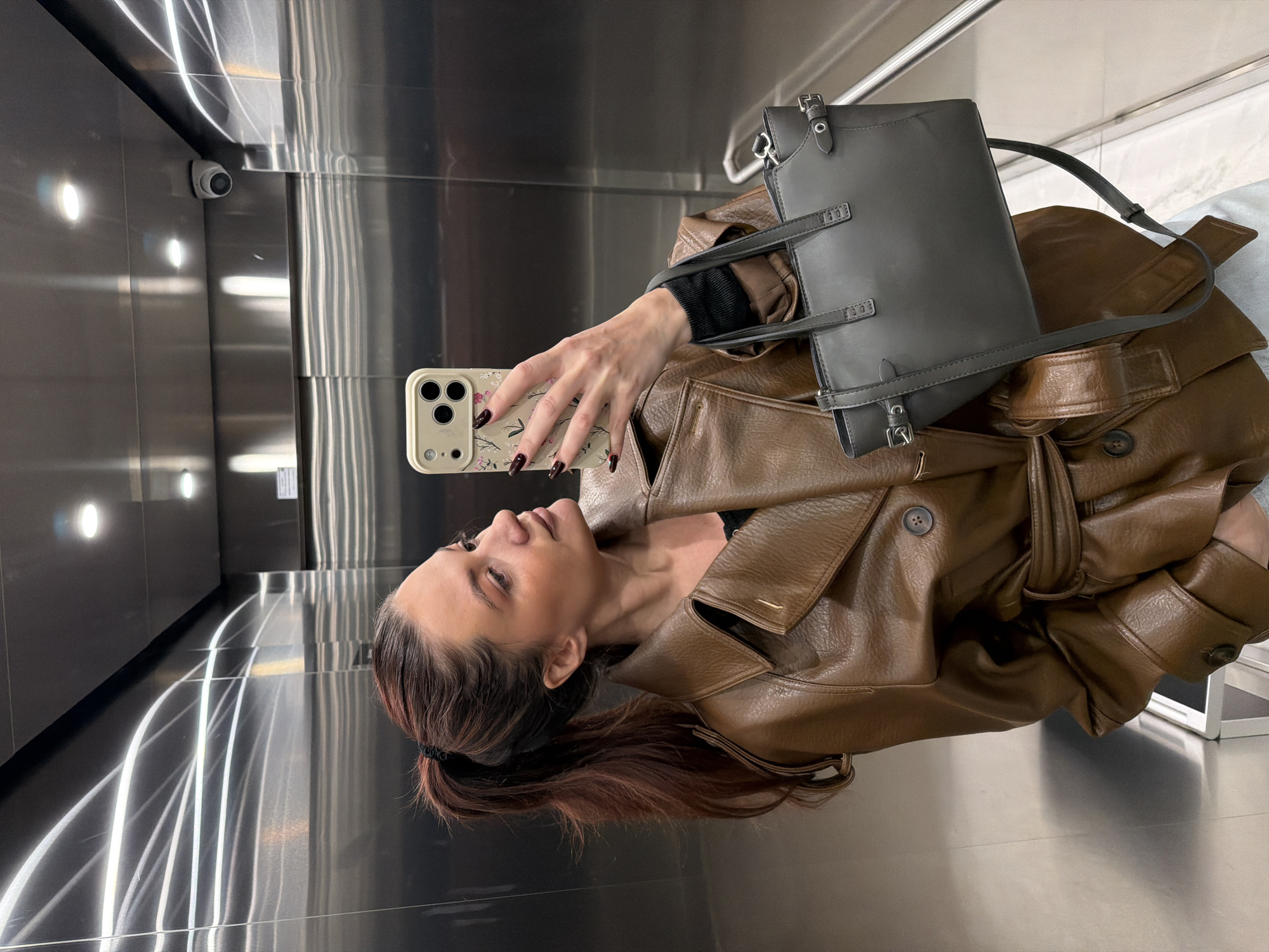 A woman with auburn hair and dark fingernails poses for a selfie in what appears to be a metal elevator or lift in Belgrade, Serbia. She is wearing a brown leather jacket and holding a phone with a patterned case. A gray handbag is slung over her shoulder. A security camera is visible in the upper left corner of the image. The interior of the elevator is illuminated with rows of lights reflected on the metal surface.