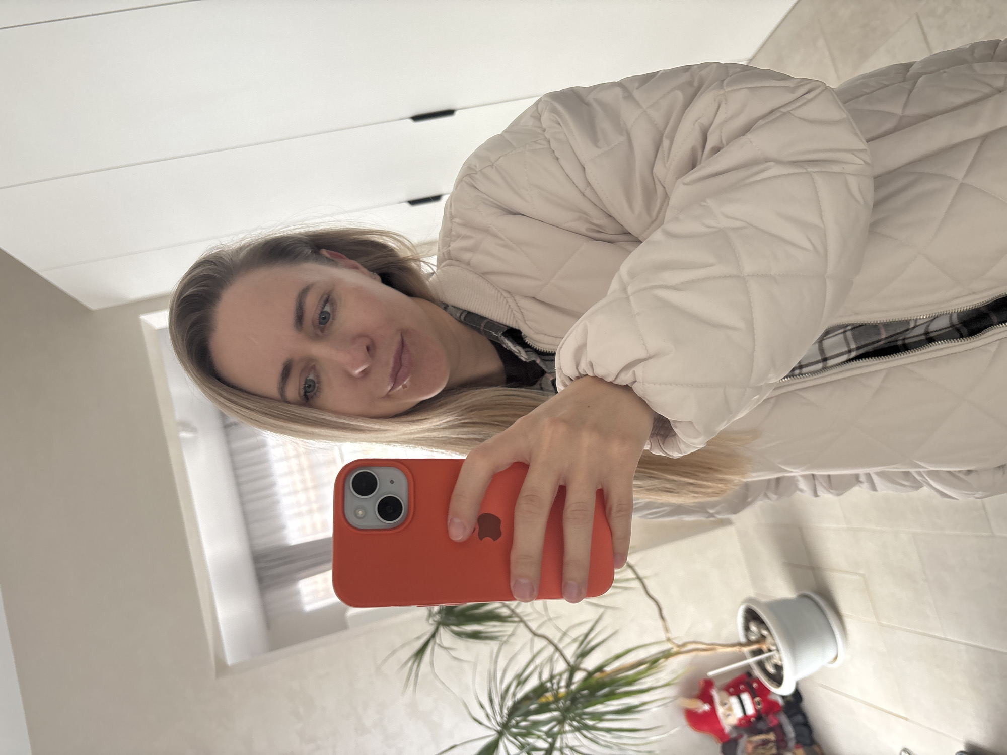 A fair-skinned woman with long blonde hair is taking a selfie in what appears to be an indoor setting. She is holding an orange-colored smartphone, and her upper body is visible, clad in a light-colored, quilted jacket with a zipper. A plaid garment appears to be worn underneath the jacket.
The background shows a white, modern wardrobe with black handles, part of a window, and what looks like a white-tiled floor. Below the camera is a potted plant, along with another figure.
There is no verifiable information to establish that this scene is located in Chaikovsky, Russia. Therefore, the location cannot be confirmed.