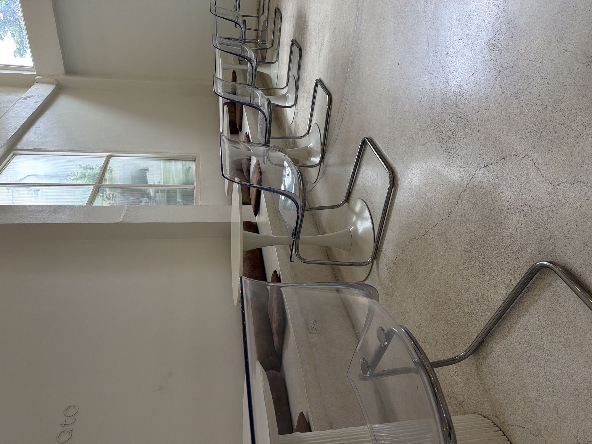 The image shows the interior of a modern cafe with a minimalist design, possibly located in the Chang Phueak area of Thailand. The cafe features white, tulip-style tables and clear acrylic chairs with chrome legs, arranged in a row along a built-in bench with brown cushions. Large windows allow natural light to illuminate the space. The walls are painted white, and the floor is a textured concrete. The overall aesthetic is clean, bright, and airy, suggesting a contemporary dining or social space.