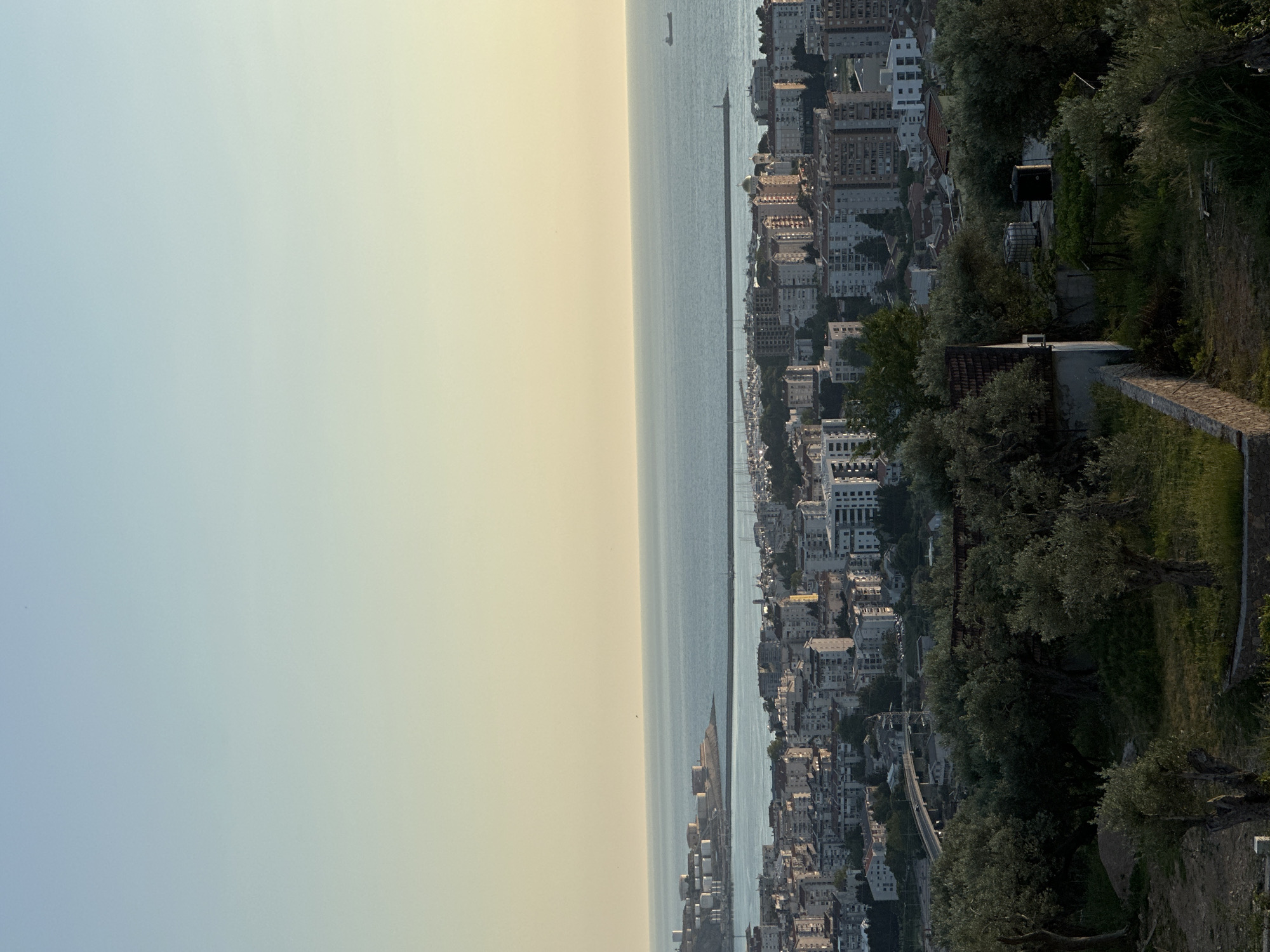 The image shows a cityscape in Velembusi, Montenegro, juxtaposed against the Adriatic Sea. The sky is a light, hazy yellow and blue gradient, indicative of sunrise or sunset. A body of water separates the sky from a densely populated urban area. The buildings are largely white or light-colored, with varying heights. Vegetation is present on the right side of the frame, closer to the viewer. At least one ship is visible on the horizon.