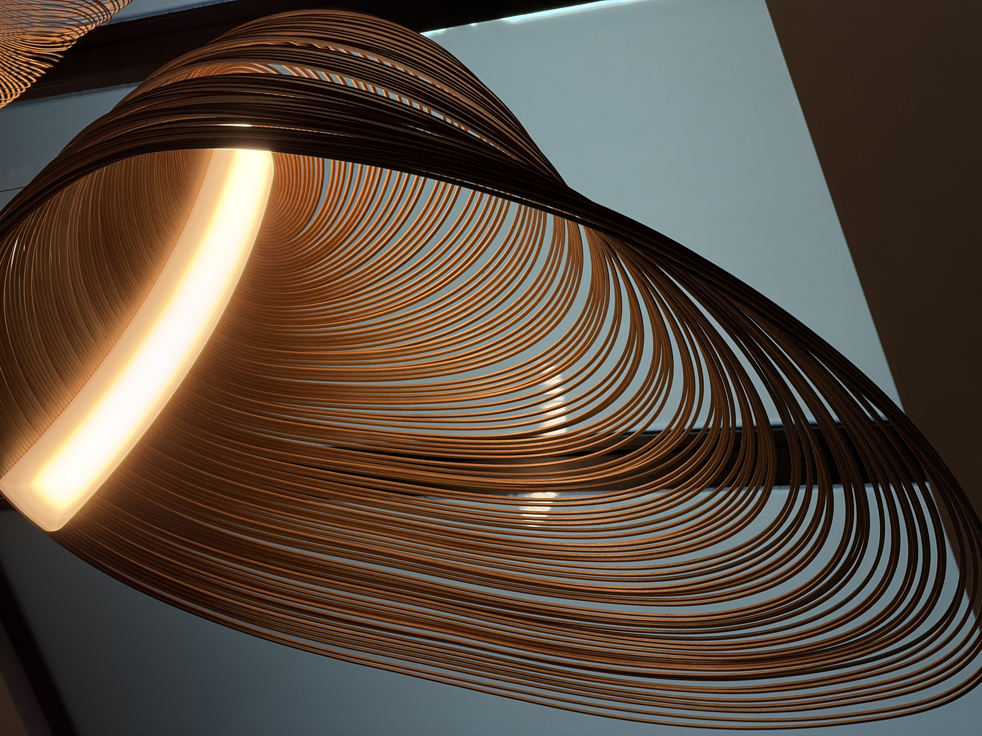 The image shows a decorative lighting fixture with a series of concentric, curved, brown bands. A linear light source is integrated within the fixture, emitting a warm, yellow glow. The background appears to be a plain, light gray surface with a dark gray element on the right. The fixture is likely indoors. The location in Kuta, Indonesia, is not apparent from the image.