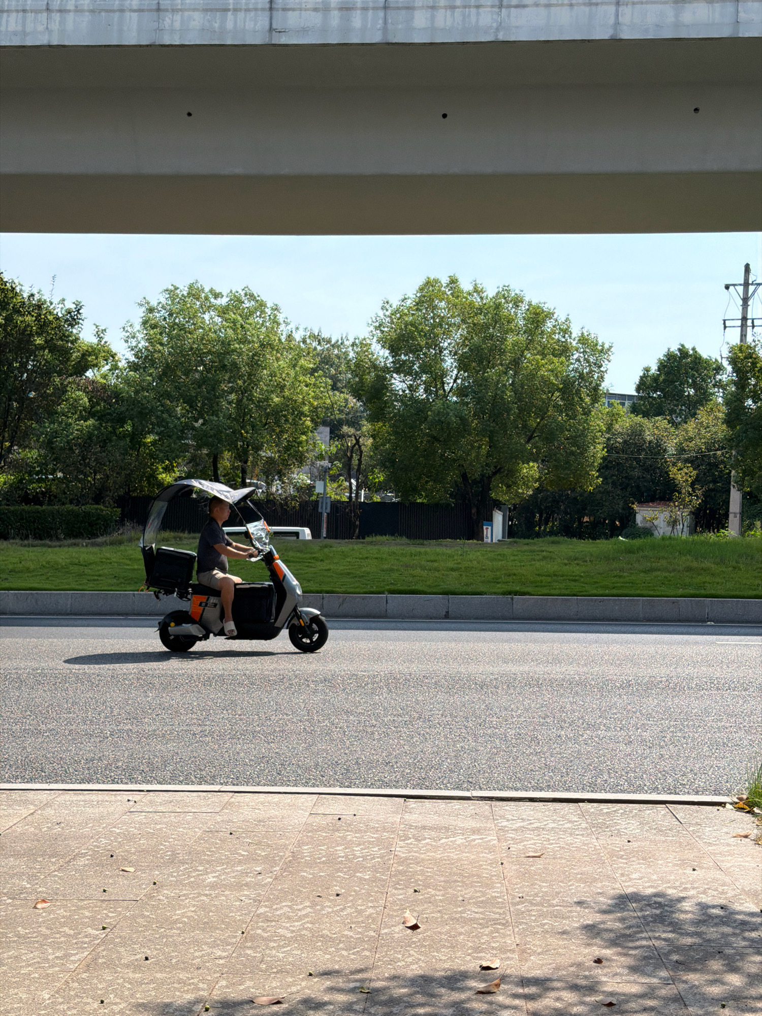 A man is riding a three-wheeled electric scooter on a paved road. The scooter has a canopy for shade and storage compartments. The man is wearing a dark t-shirt and shorts, and appears to be looking forward. The road is bordered by a grassy verge and trees. In the background, there is a concrete overpass and a utility pole. The scene appears to be in an urban or suburban area. The lighting suggests it is daytime. The area is likely located in Jiangjin, China, given the context provided.