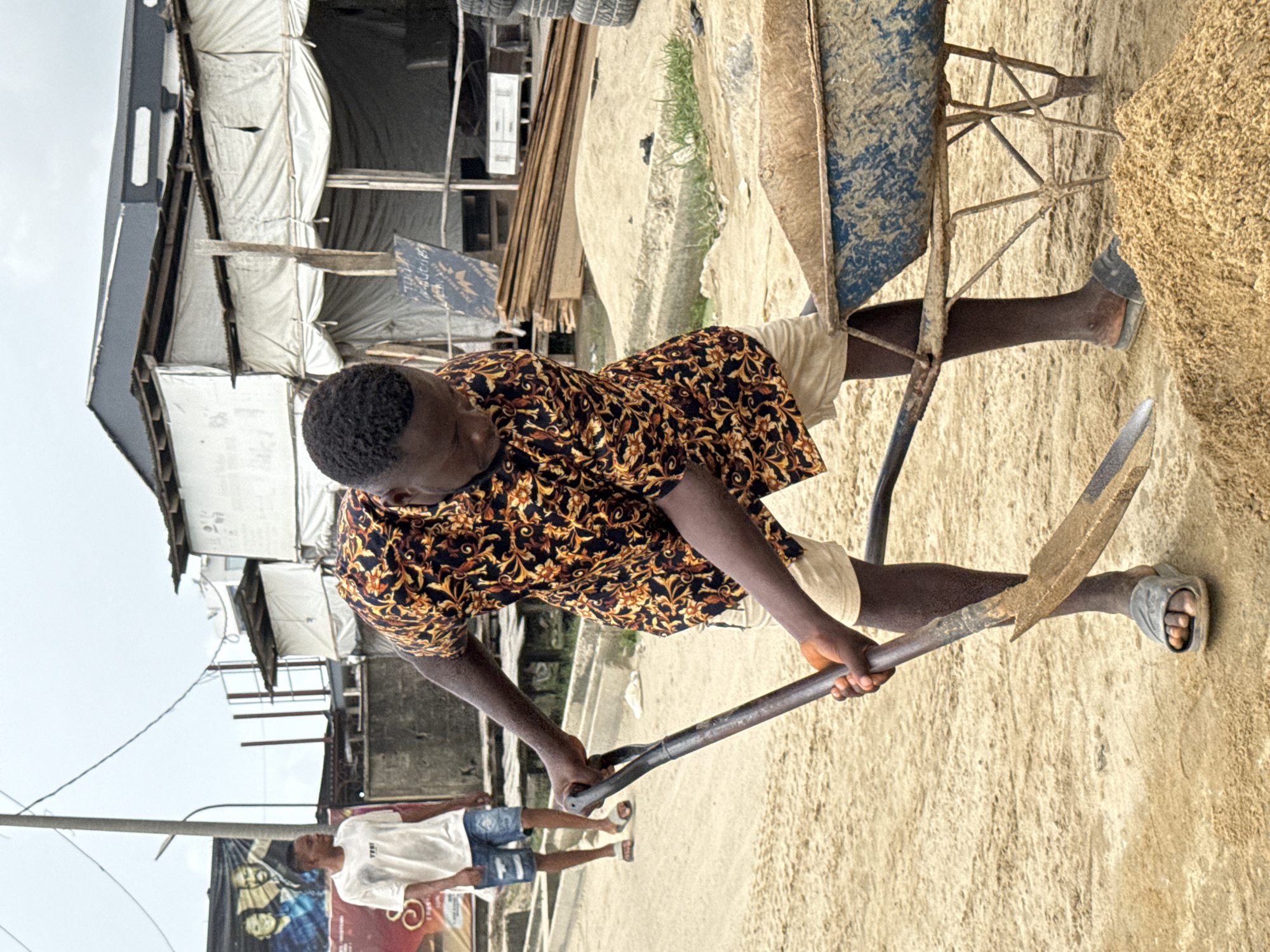 A man is shoveling gravel or sand into a wheelbarrow on a sandy or gravelly surface. He is wearing a patterned shirt, light-colored shorts, and sandals. Another man is standing in the background wearing a white shirt and jean shorts. The location appears to be in a city, possibly Ado, Nigeria, indicated by the presence of semi-permanent structures with white coverings and posters. There is exposed lumber and stacked tires visible in the background. A utility pole can be seen near the man in the background.