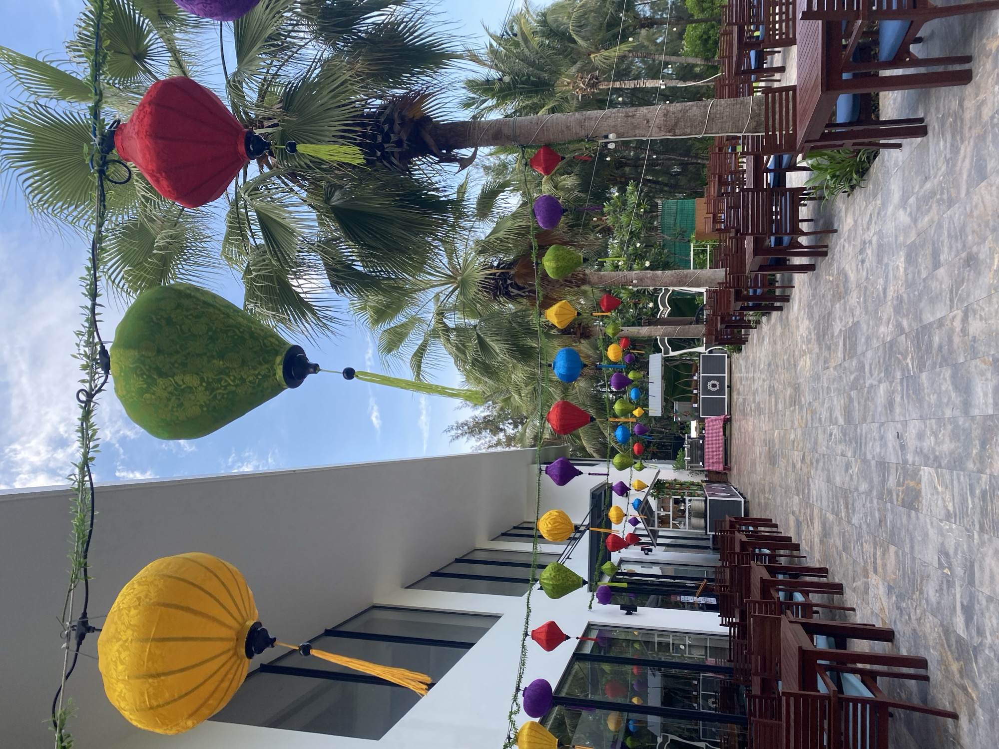 An outdoor restaurant in Bac Cam Ranh Ward, Vietnam. Rows of wooden tables and chairs line a walkway. Colorful lanterns hang between palm trees and the side of a white building. A sound system sits at the end of the walkway. The sky is blue.