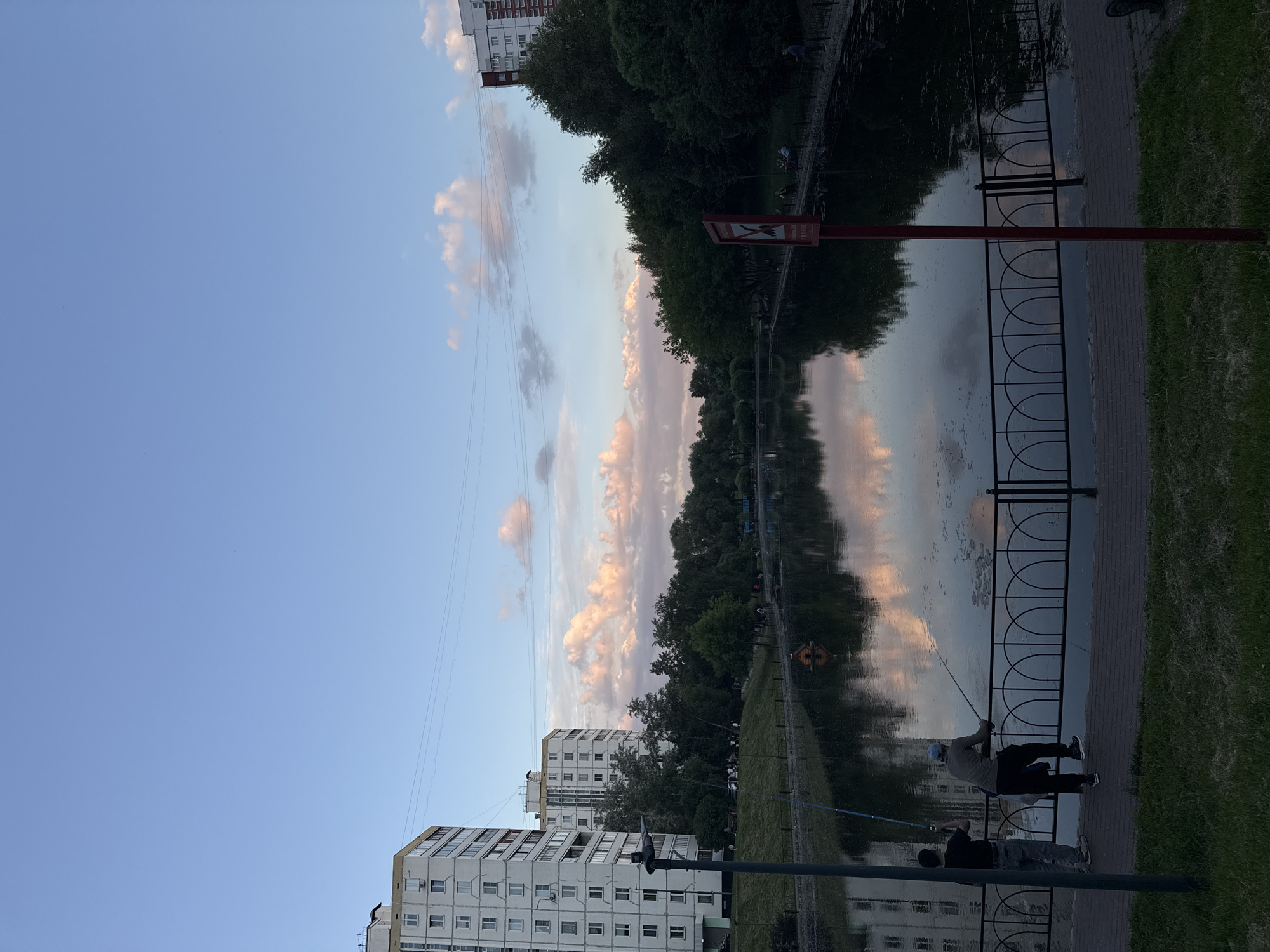 The image, correctly oriented, depicts a serene urban park scene during late afternoon or early evening. A clear blue sky with scattered cumulus clouds, tinted with soft pink and orange hues from the setting sun, dominates the upper portion. The clouds are vividly reflected in the calm waters of a canal or narrow river that stretches across the midground.
Along the water's edge in the foreground, a paved path is visible, bordered by a dark metal fence with decorative patterns and green grass. Two individuals are engaged in fishing from this bank. One person, wearing a light-colored top, dark pants, and a cap, holds a fishing rod, clearly visible against the water. The second person is partially obscured by a dark pole in the immediate foreground, also appearing to fish.
Lining the opposite bank of the water body is a dense row of green trees, their foliage mirrored in the reflective surface of the water. Towards the left background, several multi-story residential buildings, featuring numerous windows and balconies characteristic of Soviet-era architecture, rise against the sky.
A red sign with white details stands near the water's edge on the right side. It displays a white warning symbol, possibly indicating "no swimming," and unreadable white Cyrillic text below it. The overall atmosphere is peaceful and recreational, with the warm cloud colors suggesting a beautiful sunset beginning to unfold over this Moscow, Russia, setting.