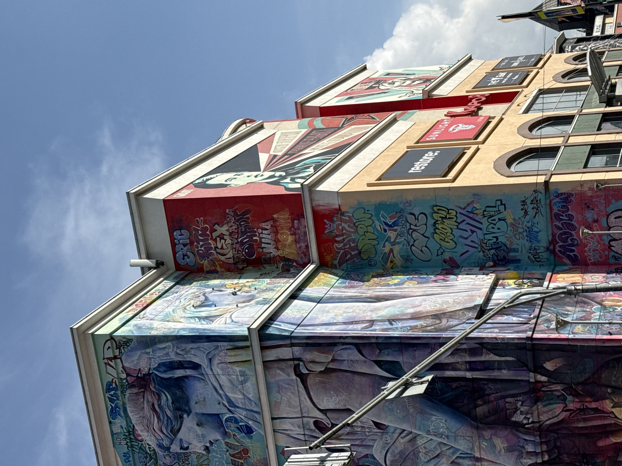 A low-angle shot depicts a multi-story building with various murals and advertisements in Moscow, Russia. The murals cover different sections of the building, displaying various artistic styles, including portraiture and abstract graffiti. The advertisements on the building include the words "restore," "Sunlight," and "The Gap." The building's architecture includes arched windows and a light-colored exterior with brown trim. The sky is blue with some white clouds. A metal pole is in the foreground.