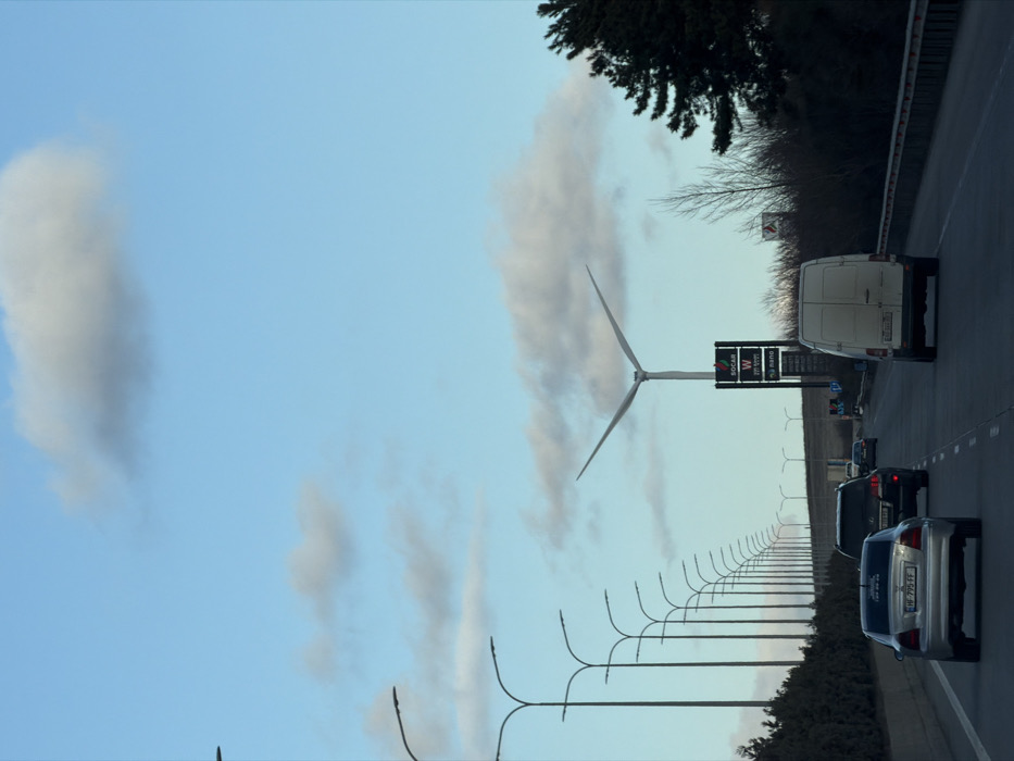 The image depicts a multi-lane highway in Ruisi, Georgia, under a blue sky with scattered white clouds. A prominent two-bladed wind turbine stands in the mid-ground on the right, positioned behind a tall black sign displaying "SOCAR", "W", and "nano" logos. A continuous line of streetlights with curved arms extends along the left side of the road, receding into the distance. Multiple vehicles are observed on the asphalt pavement. In the foreground, a silver sedan with a Georgian license plate "HF-795-FF" is in the left lane, followed by a dark SUV in the adjacent right lane. Further ahead in the right lane, a white panel van is visible from the rear. Roadside vegetation, including dense dark green bushes and some bare deciduous trees, flanks both sides of the highway, with a metal guardrail present along the right edge of the road. Distant terrain rises gently beyond the immediate roadside. No discernible people are visible.