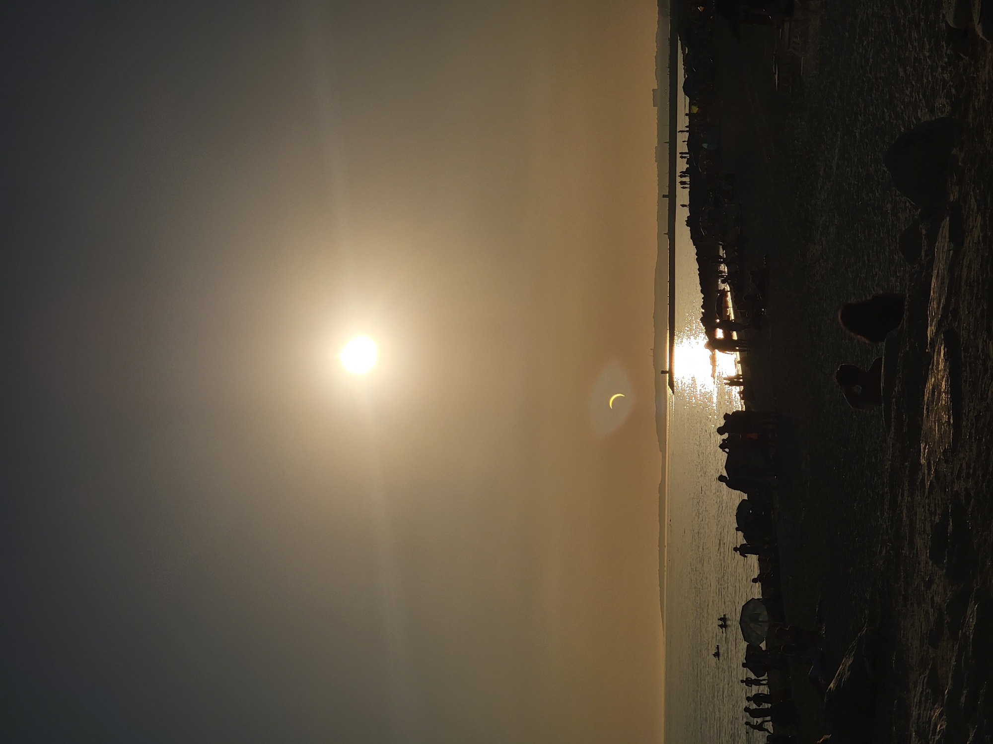 The image depicts a sunset over the Atlantic Ocean in Quarteira, Portugal. A partial solar eclipse is visible in the sky, with the sun appearing as a bright orb partially obscured by a darker object, creating a crescent shape. The sun's reflection shimmers on the water. Silhouetted figures of people are scattered across a sandy beach and along a rocky breakwater. Some individuals are wading in the shallow water, while others are gathered on the shore. Beach umbrellas and tents are visible amongst the crowd. The overall atmosphere suggests a late afternoon or early evening gathering at the beach.