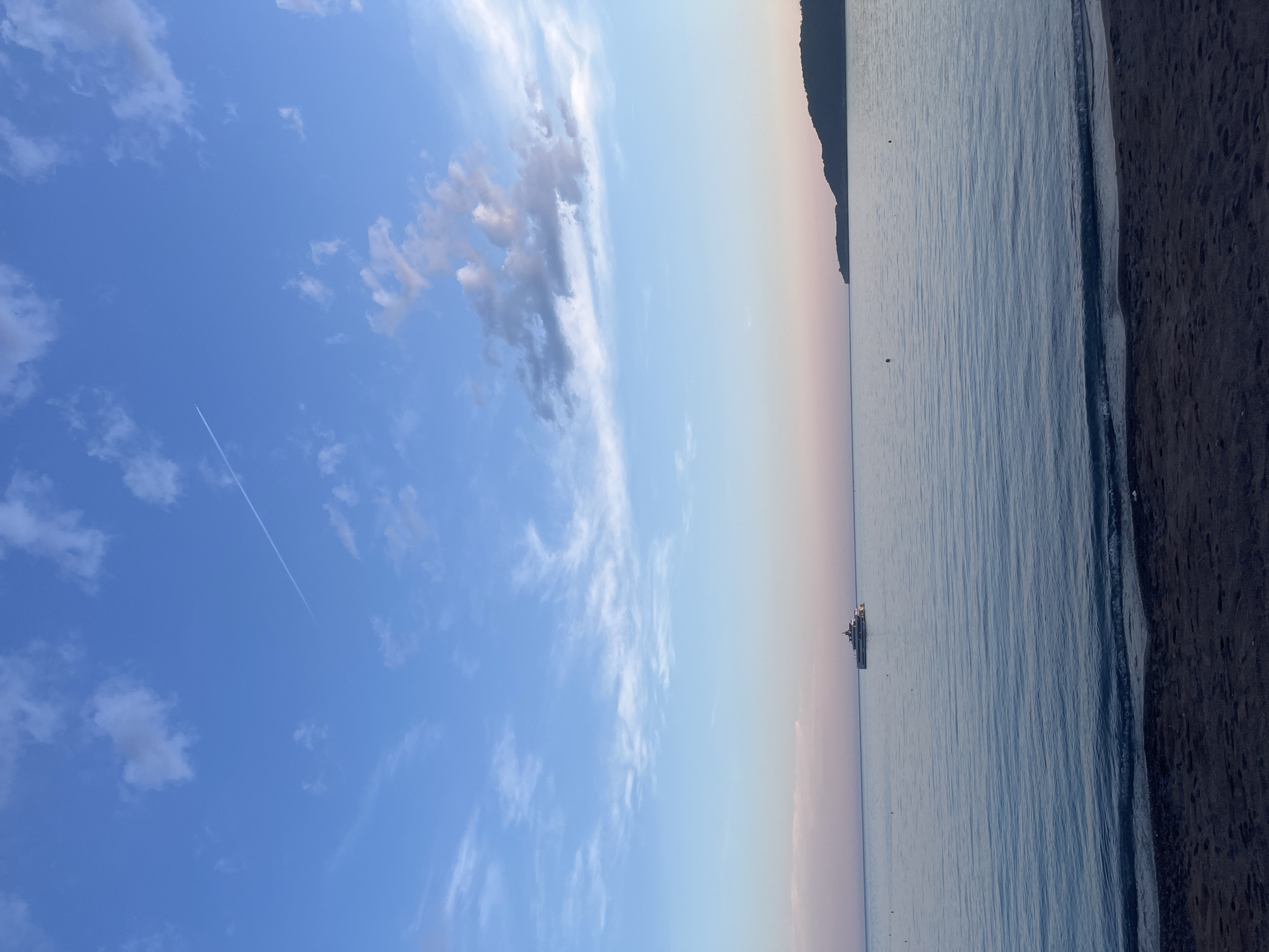 The image shows a beach scene in Bečići, Montenegro. The view is of the sky, the Adriatic Sea, and the coastline. In the sky, there are scattered clouds, including a notable cluster, and a contrail. The sea is calm with gentle ripples, and the beach is composed of dark sand and small pebbles. A cruise ship or large vessel is visible on the water near the horizon. The coastline includes a headland and the edge of the beach.