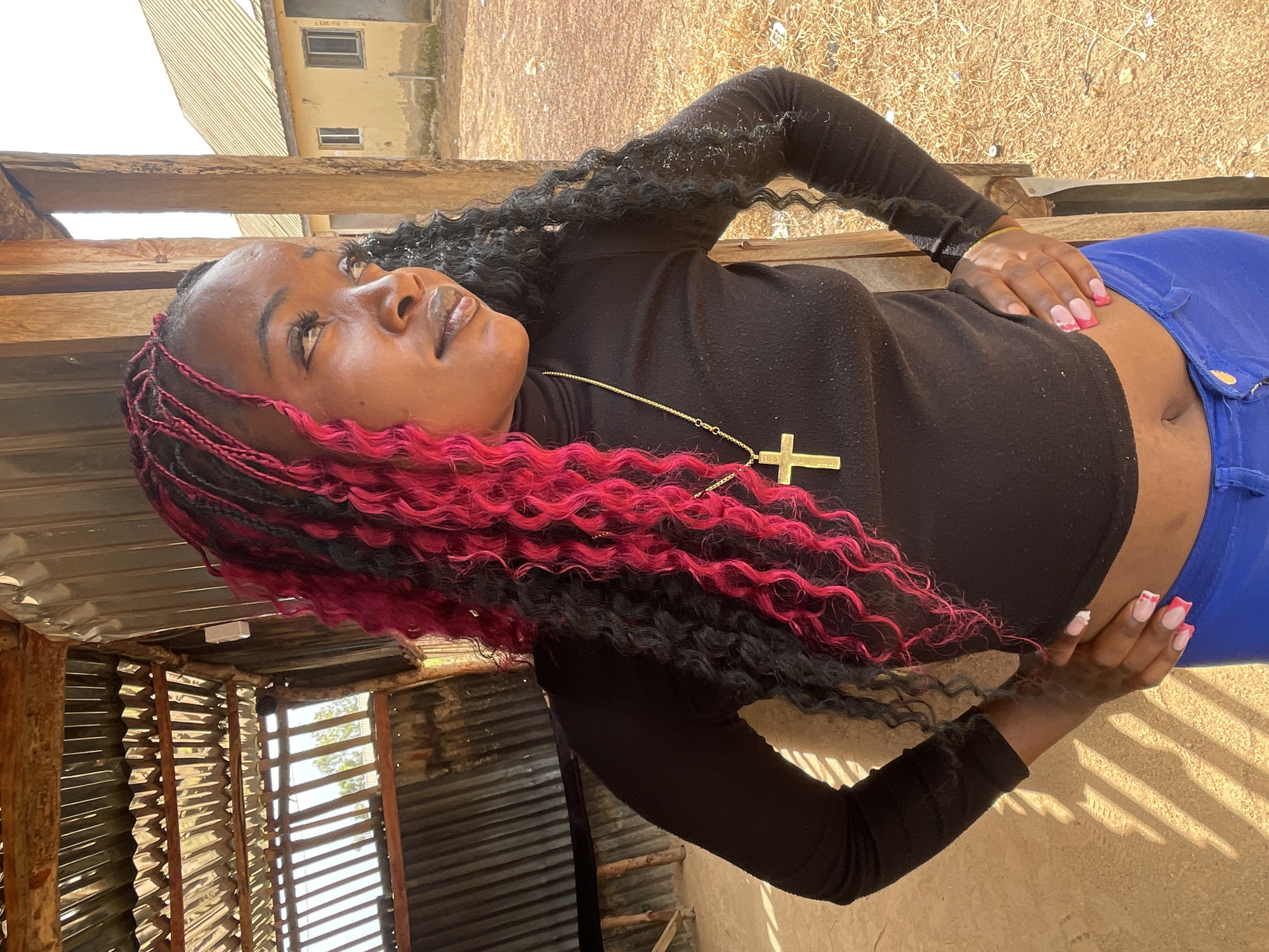 In Durumi, Nigeria, a young Black woman with braided hair, partially dyed bright pink, stands outdoors. She is wearing a black turtleneck shirt and vibrant blue pants. A gold cross necklace adorns her neck. Her hands are placed on her hips, and her fingernails are painted with a pink and white French manicure. The background reveals a rustic structure with a corrugated metal roof and wooden supports, suggesting a simple, perhaps unfinished, dwelling. Beyond this structure, two yellow buildings with light-colored roofs are visible, set against a dry, earthy landscape with sparse vegetation. The lighting suggests it is daytime, with shadows casting diagonal patterns on the ground.