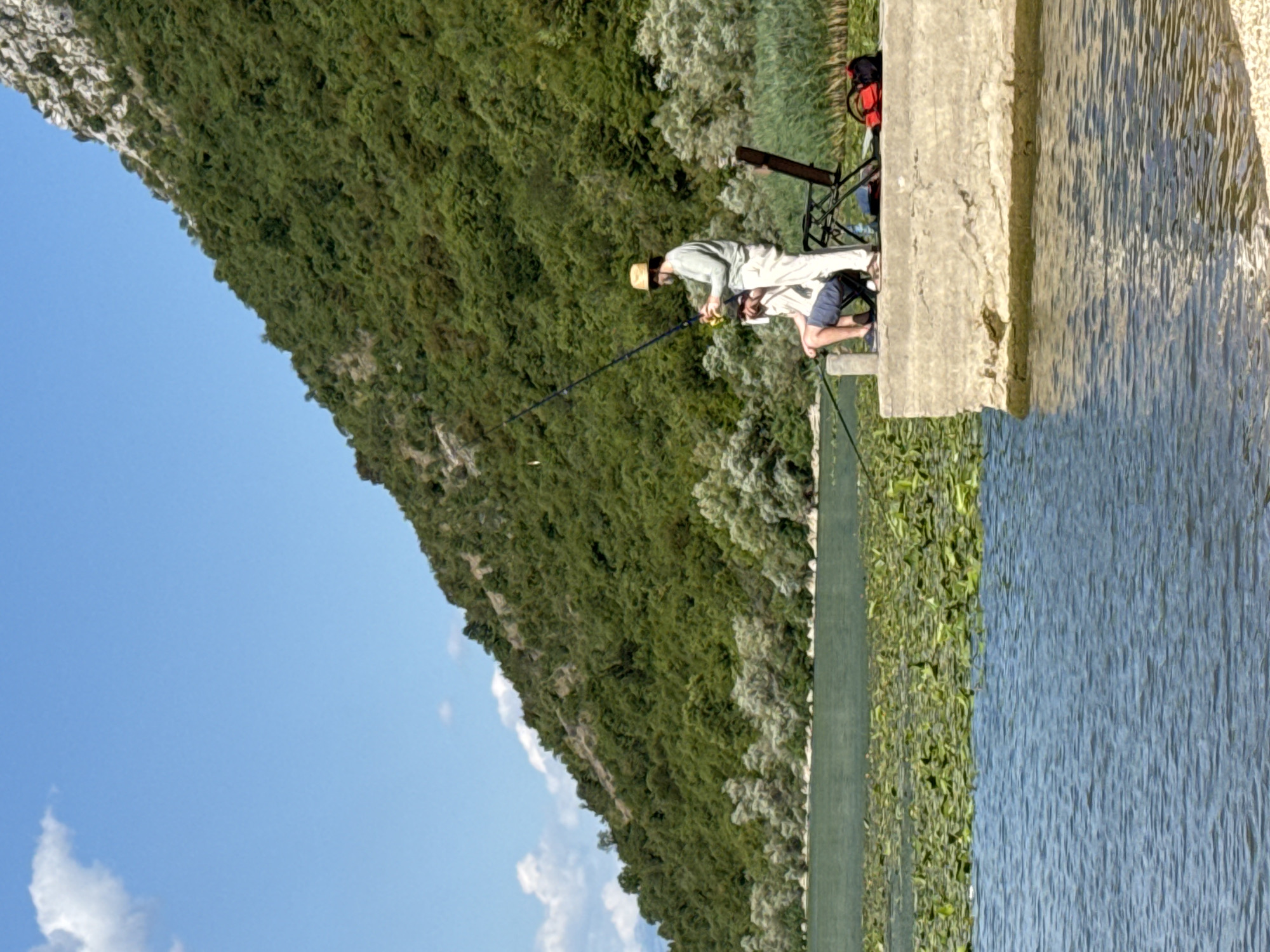 The image depicts a serene outdoor scene, likely in Seoca, Montenegro, focusing on two individuals engaged in fishing on a concrete pier. The foreground is dominated by the calm, blue water of a lake or river, with a dense patch of lily pads visible on the surface. To the right, a concrete pier juts out into the water. On the pier, two people are fishing. One, wearing a light-colored long-sleeved shirt, light-colored pants, and a straw hat, stands holding a fishing rod. The second person is seated, wearing a light shirt and dark shorts, also holding a fishing rod. A folded black chair is positioned behind the standing individual, and a red and black backpack rests on the pier near the chair. In the background, a lush, green, tree-covered mountain slope rises steeply, bathed in sunlight under a clear blue sky with a few wispy clouds. The overall impression is one of peaceful recreation in a picturesque natural setting.