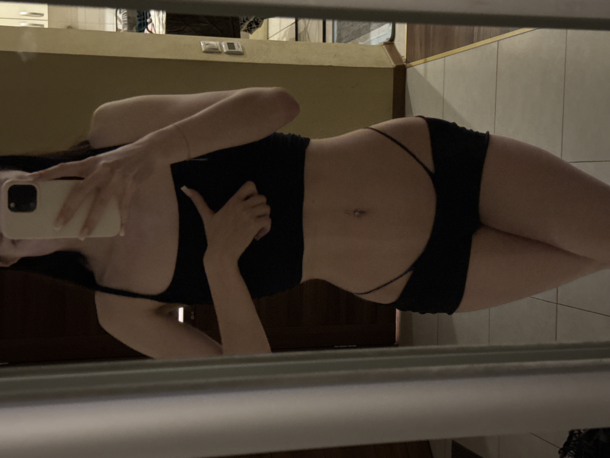 A young woman is taking a mirror selfie in what appears to be an apartment hallway. She is wearing a black crop top and black underwear. Her left hand is touching her chest while her right hand is holding a smartphone, which is reflecting in the mirror. The background shows a wooden door, a yellow-painted wall with light switches, and a tiled floor. The overall lighting is dim, suggesting it's indoors. There is no visual information to confirm the location as Khimki, Russia.