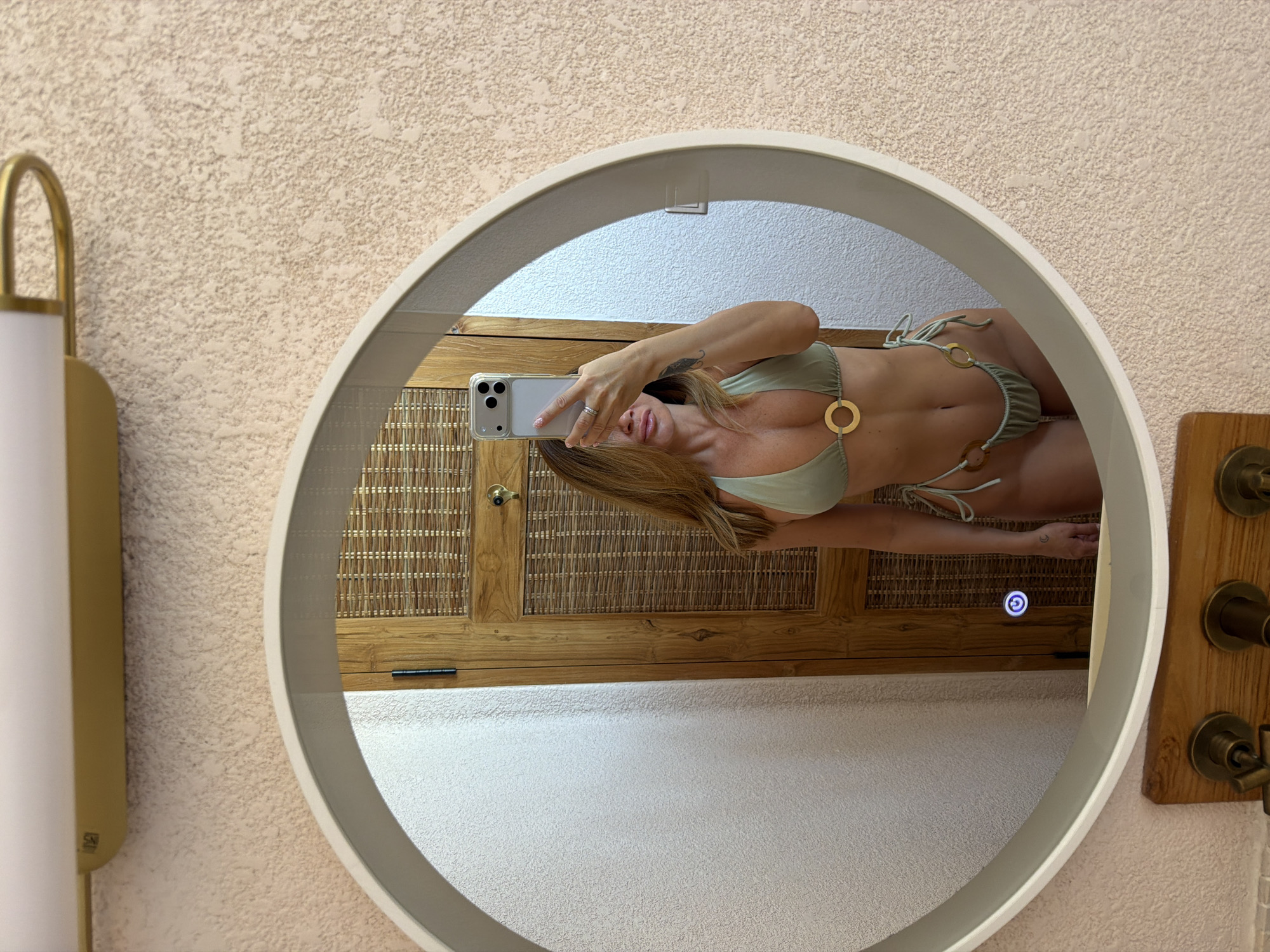 This rotated image captures a mirror selfie taken by a person, likely female, in what appears to be a bathroom or dressing area. The subject, visible in the reflection of a large, white-framed circular mirror with an integrated light, is wearing a light sage green two-piece bikini adorned with gold-colored rings. She holds a silver smartphone (an iPhone) up to take the photo, partially obscuring her face.
The setting visible in the reflection features wooden elements, including a cabinet or door with a woven rattan panel and a brass-colored knob, suggesting a natural and possibly tropical aesthetic. On the right, a wooden panel with multiple brass-colored knobs or spigots is reflected, indicative of a sink or shower fixture. The walls surrounding the mirror have a textured, light beige finish. On the left side of the image, a wall-mounted light fixture with a gold-colored arch and a white cylindrical shade is partially visible, bearing the text "SXI". The mirror itself has a touch-sensitive power button, glowing blue with a "G" symbol, suggesting it is illuminated.
The overall scene, with its natural materials and a person in swimwear, aligns with the provided context of Mengwi, Indonesia, hinting at a resort or hotel environment. The activity is a casual mirror selfie, with no strong emotions discernible.