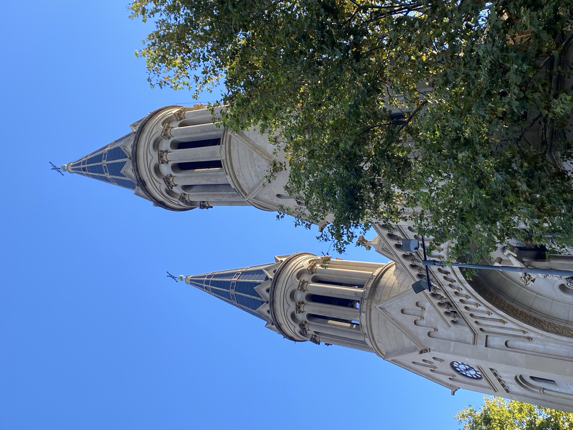 The image features a low-angle view of two ornate towers against a clear blue sky, partially obscured by foliage. The towers are part of the Basilica del Santísimo Sacramento, located in Buenos Aires, Argentina. The towers are constructed of light-colored stone and feature pointed spires with dark, tiled roofs. Architectural details include columns, arches, and decorative carvings. One of the towers has a clock face visible near its base. A streetlight is seen attached to the base of one of the towers.