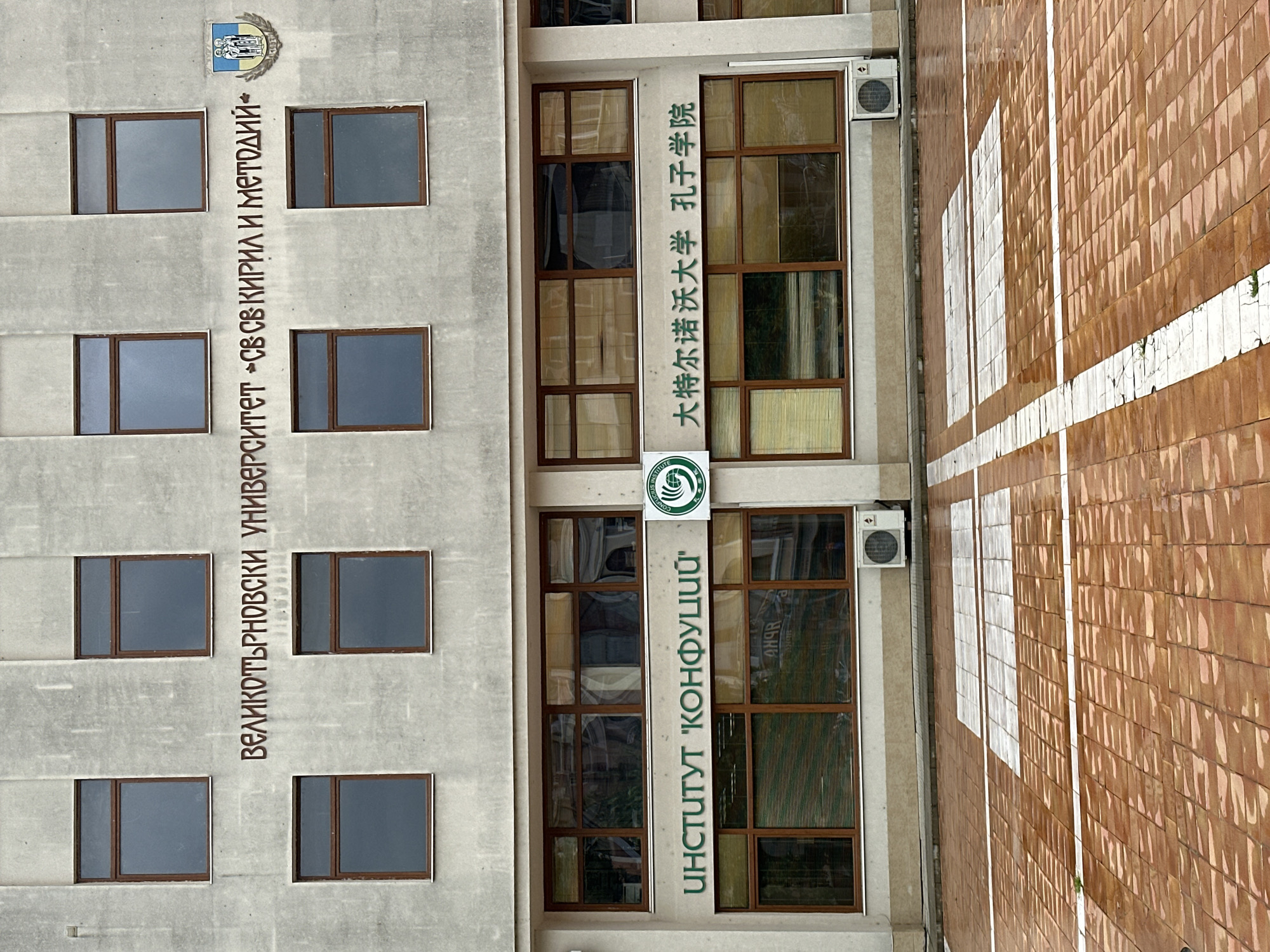 The image shows the exterior of a building in Veliko Tarnovo, Bulgaria. Two sections of the building are visible. On the left, in brown lettering, the Bulgarian text "ВЕЛИКОТЪРНОВСКИ УНИВЕРСИТЕТ СВ. СВ. КИРИЛ И МЕТОДИЙ" is vertically aligned. To the right, another section features the text "ИНСТИТУТ 'КОНФУЦИЙ'" in green, as well as Chinese text above the logo of the Confucius Institute. Air conditioning units are mounted near the windows. A red-tiled surface with white lines is seen on the right.
