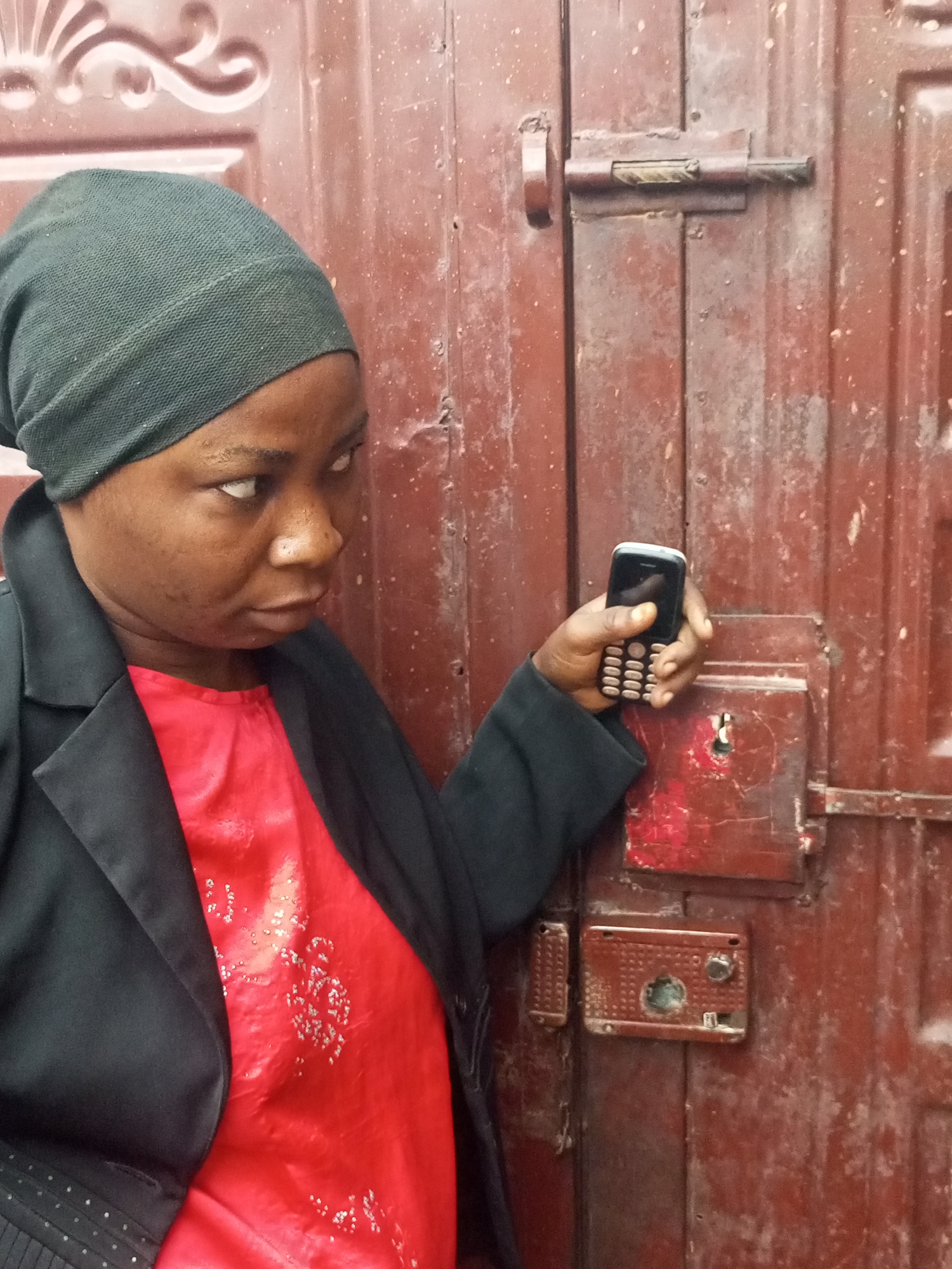 An African woman wearing a dark green headscarf and a black jacket over a red shirt stands in front of a weathered, reddish-brown metal door. She is holding a small, black mobile phone in her right hand, angled towards a lock mechanism on the door. The door has multiple panels and appears to be secured by two latches, one of which is partially visible in the upper left. The lock mechanism to the right of the door is a rectangular metal plate with a keyhole and a knob. The woman's face is turned to her left, and her gaze is focused on the lock. The lighting suggests it is daytime, but the environment is somewhat dim. The location is identified as Tempure, Nigeria.