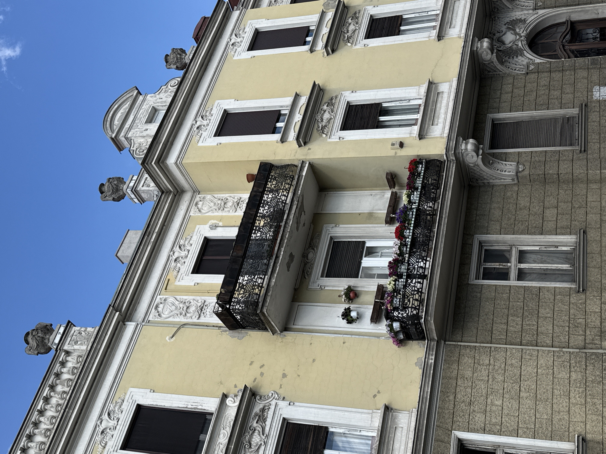 This low-angle photograph showcases the ornate facade of an old building in Belgrade, Serbia, under a bright blue sky with a few wispy clouds. The image is tilted, making the building appear to lean towards the right.
The primary section of the building, occupying most of the frame, features a creamy yellow facade adorned with extensive white stucco decorations. These intricate embellishments include detailed moldings, cornices, and window surrounds with floral, shell, and scroll patterns, though some show signs of wear and age. Multiple windows with white frames are visible, fitted with dark brown external roller blinds or shutters, some partially open.
Two prominent wrought-iron balconies extend from the yellow facade. Both feature elaborate, lacy metalwork and are decorated with colorful potted flowers in shades of red, purple, and white, adding a vibrant touch. Along the roofline, several decorative, gargoyle-like sculptures further enhance the building's historical and ornate character.
To the right and lower part of the frame, an adjacent or lower section of the building is visible. This part has a darker grey, textured facade, possibly a roughcast finish or with a patterned relief. Its windows are simpler, some with dark shutters, while one features a multi-pane white frame. A light-colored conduit runs horizontally across this grey section.
The clear sky and bright lighting indicate it is daytime. There are no people visible, and no text is discernible on the building's exterior. The photograph captures the architectural detail and character of a historic urban structure.