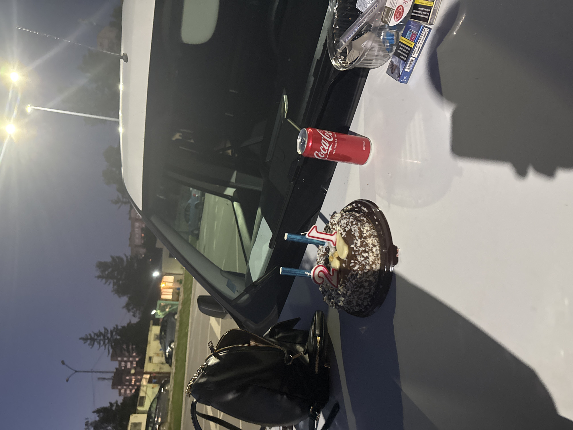 On the hood of a white vehicle parked in Unknown, Unknown, sits a black purse, a chocolate cake with number 21 candles, a can of Coca-Cola, a plastic container with plastic utensils, and two packs of cigarettes. The background includes street lights, buildings, trees, and other parked cars. It is twilight.