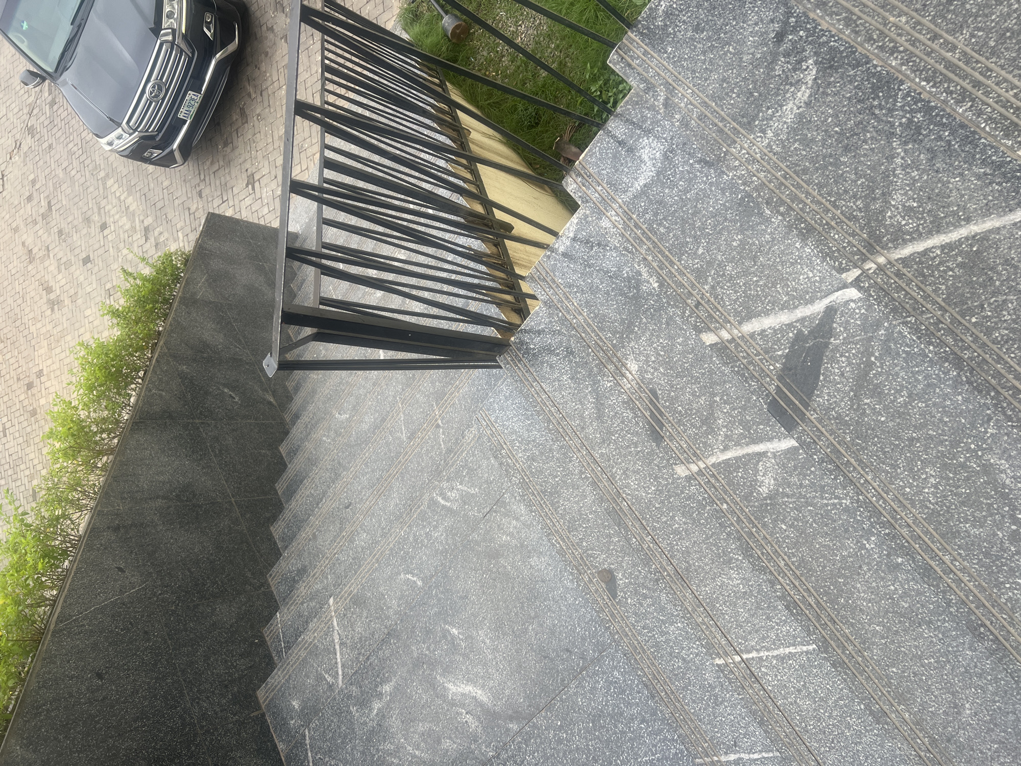 The image shows a perspective view of an outdoor staircase made of speckled grey granite. The staircase has multiple steps, each with three parallel metal strips for grip. A black metal railing with vertical bars runs along the right side of the stairs. Above the staircase, a low wall clad in darker granite is visible, topped with a row of green leafy plants. Beyond the wall, a paved area made of interlocking grey bricks is visible, with a dark-colored SUV parked on it. The SUV has a license plate that reads "AAA-923FA". The scene appears to be in an urban setting, likely within the city of Abuja, Nigeria, given the presence of the vehicle and typical landscaping.