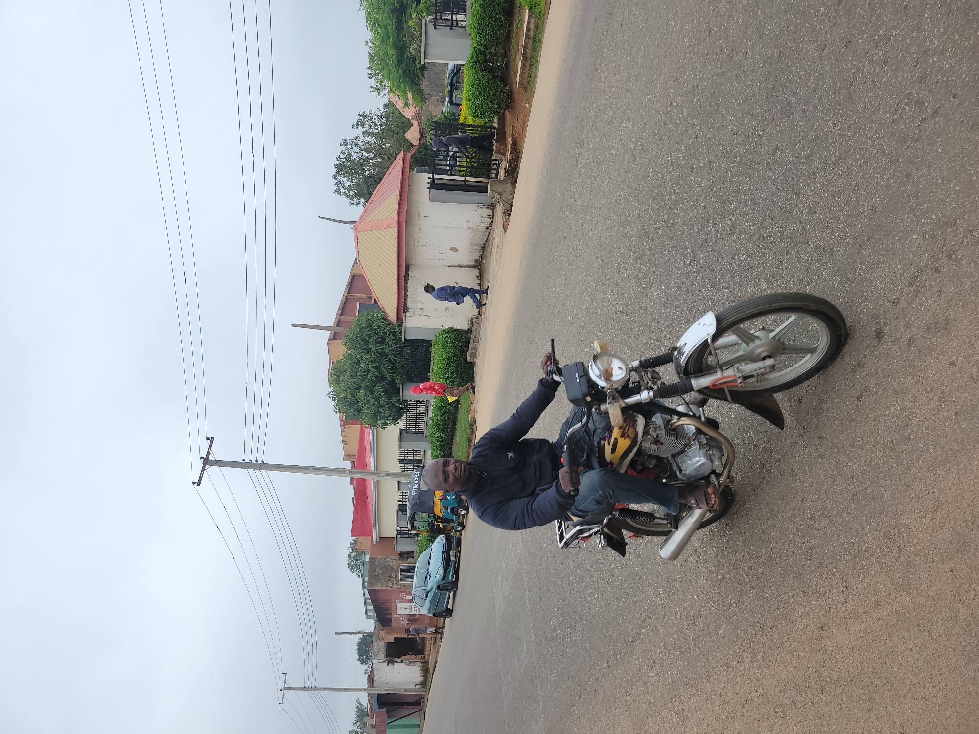 A man is riding a motorcycle on a paved road in the city of Jos, Nigeria. The man is wearing a dark blue jacket and jeans, and he is looking ahead. The motorcycle is yellow and silver, and it has a black seat. In the background, there are several buildings and trees. The sky is overcast. There are also a few other people and vehicles visible in the background. The road appears to be a residential or commercial area.