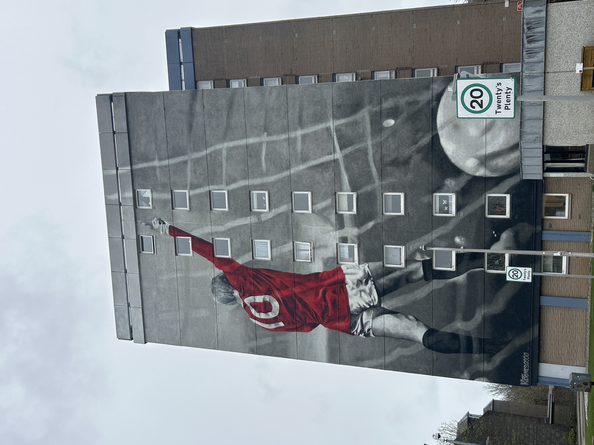 The image depicts a mural on the side of a building, likely in Aberdeen City, United Kingdom. The mural is a black and white image of a soccer player diving to head a ball, wearing a red jersey with the number 10 on the back. The building is a tall, rectangular structure with multiple rows of windows. Above the mural is another part of the building with fewer windows. To the right of the mural, there's a traffic sign that reads "20 Twenty's Plenty," indicating a speed limit. The sky appears overcast, suggesting a cloudy day. The mural has the signature "Keionesgoo" and the year "2000" written in the bottom right corner.