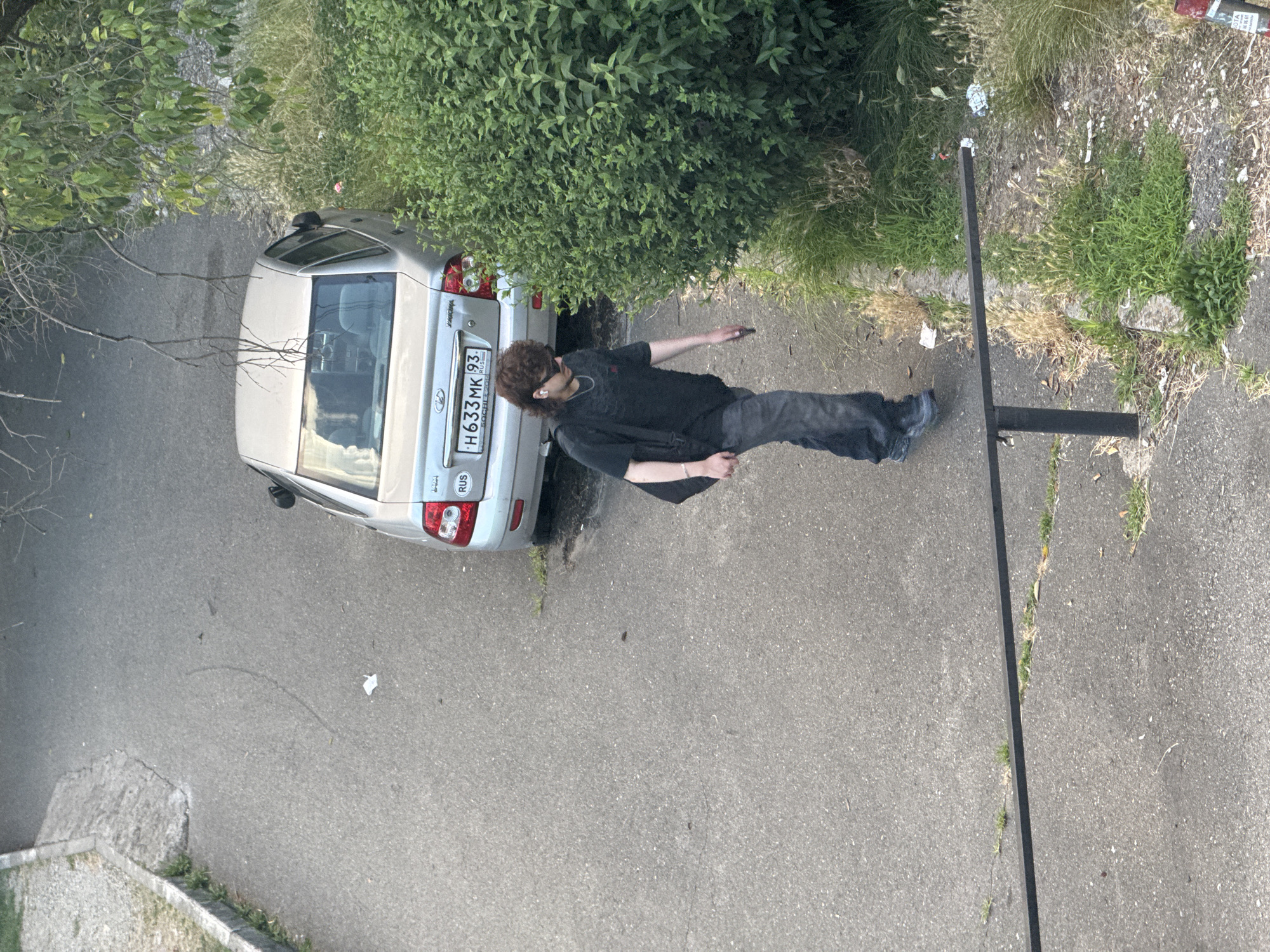 This media file shows a person standing next to a silver car. The person is wearing dark clothing and sunglasses and has curly brown hair. They are holding a mobile phone. The car is parked on an asphalt surface. In the background, there is dense green foliage and grass. A black pole is visible to the right of the scene.
The scene appears to be outdoors, with natural light suggesting it could be daytime. The presence of green vegetation and the absence of heavy coats imply mild weather. The license plate of the car reads "H633MK 93". The visible text "RUS" on the license plate indicates the vehicle is registered in Russia. Based on the provided location context of Sochi, Russia, the scene is likely set in an urban or suburban area of this city. The person's posture and the presence of a mobile phone suggest a casual moment, possibly involving communication or a brief pause.
