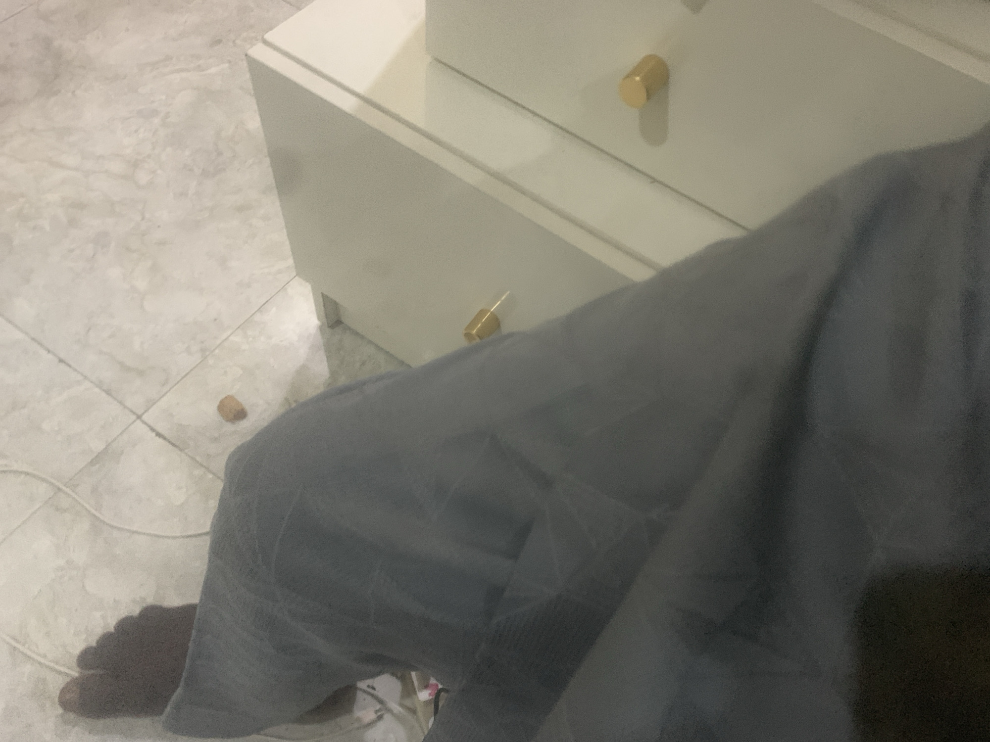 An elevated shot shows a person's legs and feet from the knees down. The individual is wearing gray pants and is seated on a tiled floor. To the right of the person are two drawers with gold knobs. The background is a light-colored tile floor. A white cord is visible on the floor near the person's feet. The location is Kuson, Nigeria.