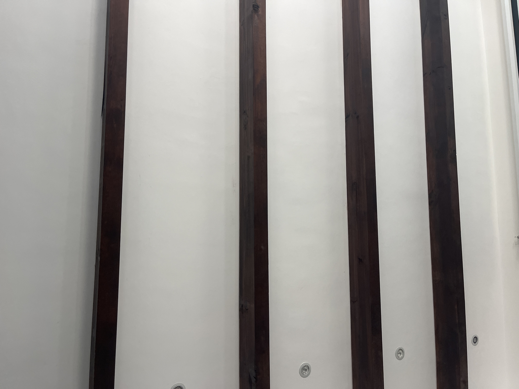 The image shows a section of a ceiling with dark brown wooden beams spaced evenly apart. The ceiling between the beams is white and features recessed lighting fixtures. The location is San Bernardino Tlaxcalancingo, Mexico.
