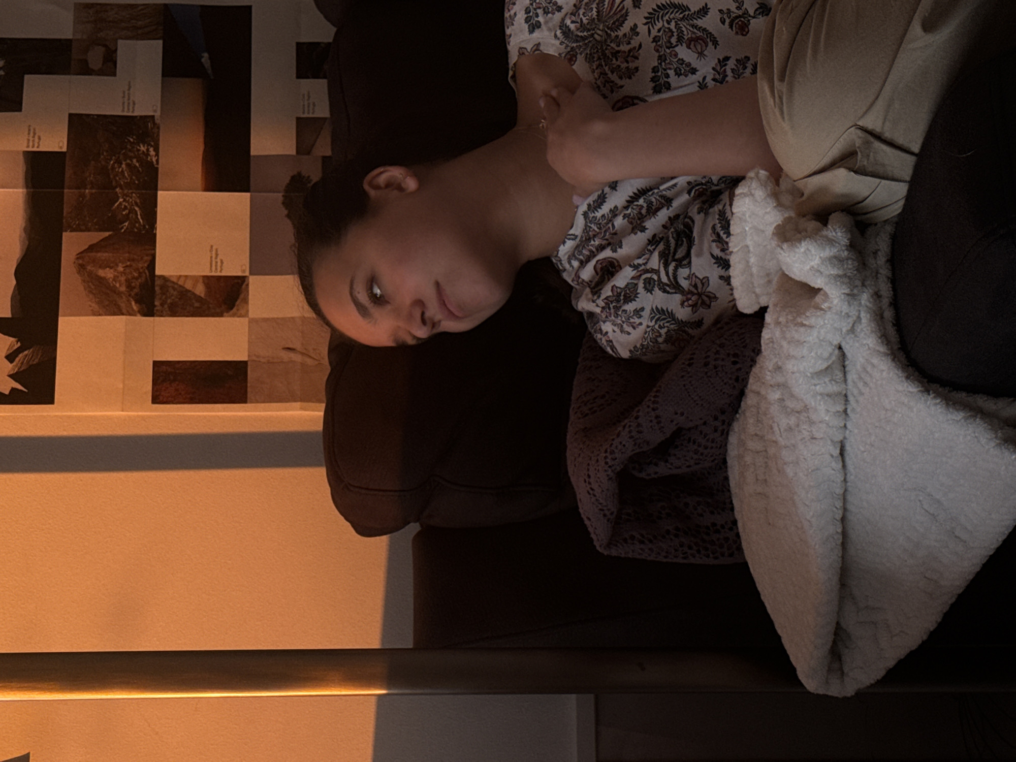 A woman is reclining on a dark-colored couch, draped with a white, textured blanket and a gray lace throw. She is wearing a patterned shirt and tan pants. The setting is indoors, near a wall painted a light color with a horizontal metal bar attached, suggesting a modern interior. A patchwork of papers with images and text is mounted on the adjacent wall. The image provides no information to determine if the photo was taken in Milan, Italy.