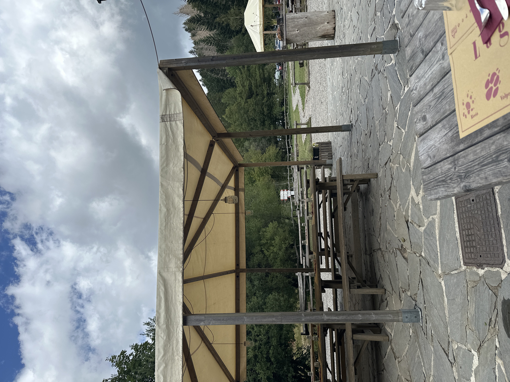 The image depicts an outdoor seating area, likely a restaurant or cafe, in Corniglio, Italy. The scene is dominated by wooden picnic tables and benches set on a stone-tiled patio. A large, light-colored canvas awning supported by wooden posts provides shade over some of the tables. In the background, there is a dense forest of green trees, and a gravel path winds through the area. Several parked cars are visible in the distance. The sky is partly cloudy. There are no people visible in the image. The overall impression is one of a rustic, outdoor dining setting in a natural environment.