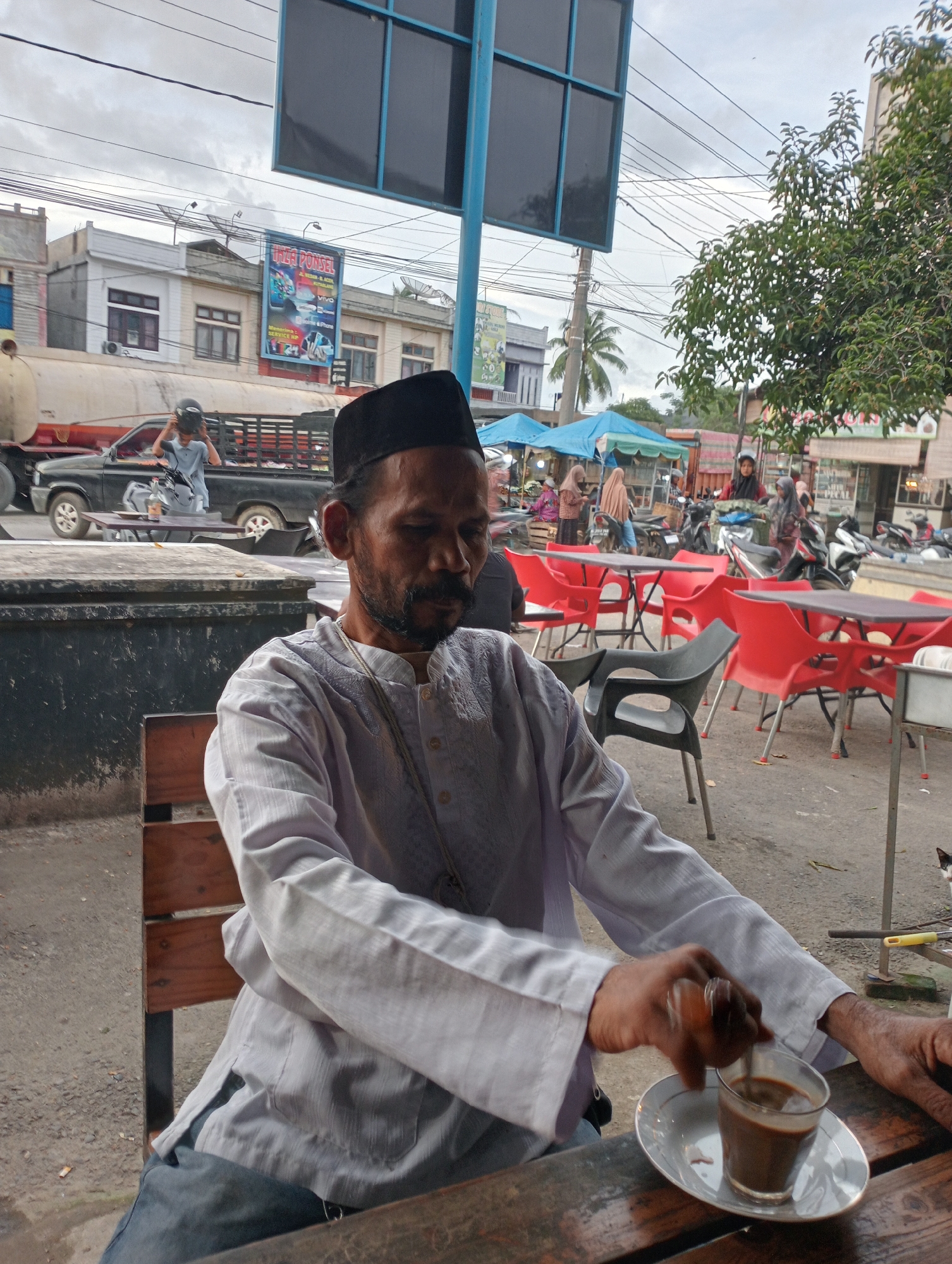 The image captures a man seated outdoors in Tingkeummanyang, Indonesia, presumably at a cafe. He's stirring a small glass of coffee. The man is wearing a black peci hat and a light-colored, long-sleeved shirt. His posture is relaxed. In the background, there's a street scene with passing vehicles, including a pickup truck with a rider. Several red chairs and tables are visible, along with people moving about, suggesting a public or commercial space. Utility poles and overhead wires are present, indicative of a built-up environment. A sign for "UZA PONSEL" is visible on a building in the background. The overall setting appears to be a typical street scene in an urban area.