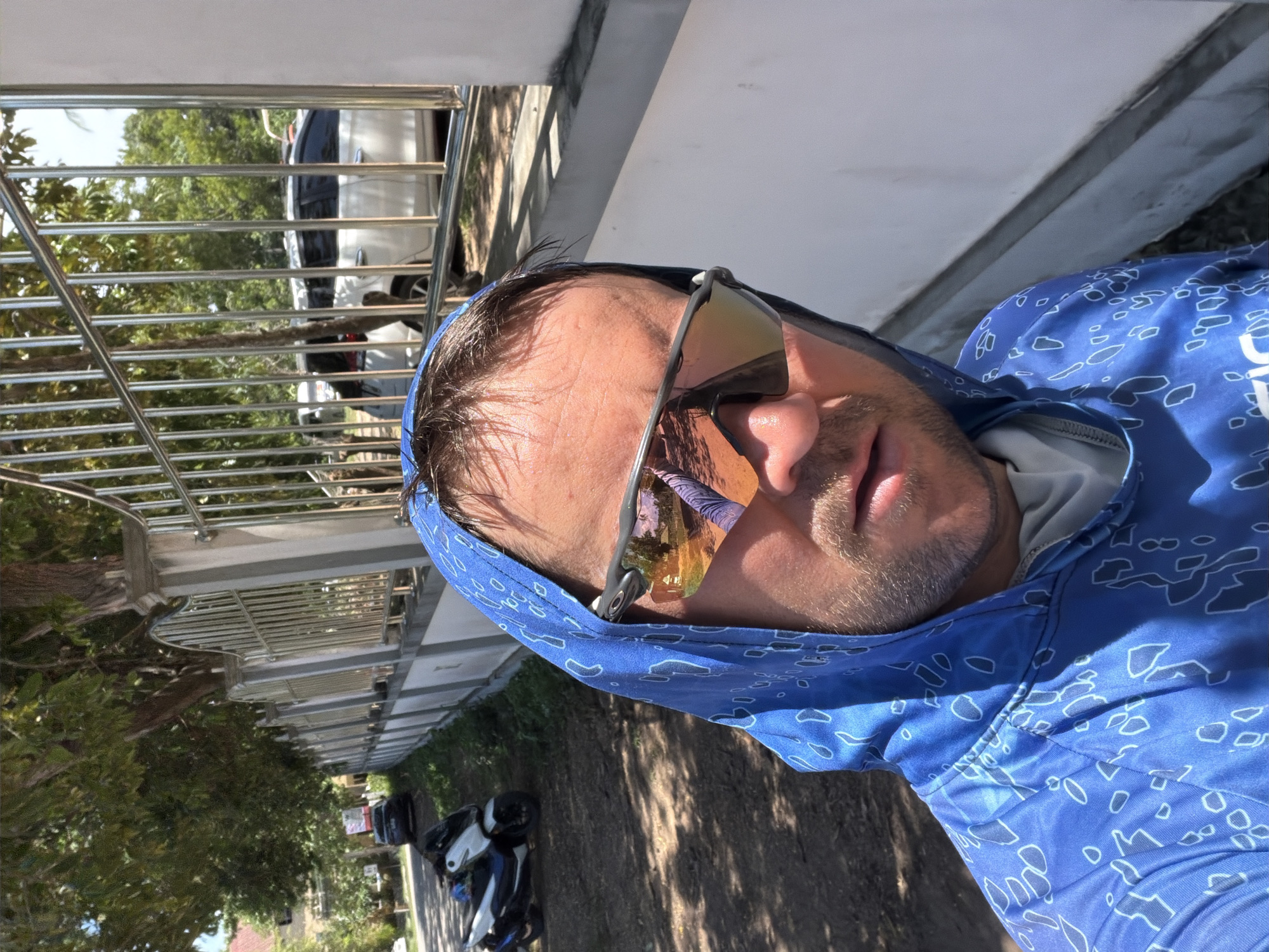 A man wearing a blue hooded jacket with a white pattern and reflective sunglasses is taking a selfie outdoors in Baan Bo Phut, Thailand. The man's face, partially obscured by the hood and sunglasses, is illuminated by sunlight. The background shows a street scene with a concrete wall and a metal fence, beyond which are trees and a white car. A scooter is parked on the dirt road to the left. The environment suggests a bright, sunny day.