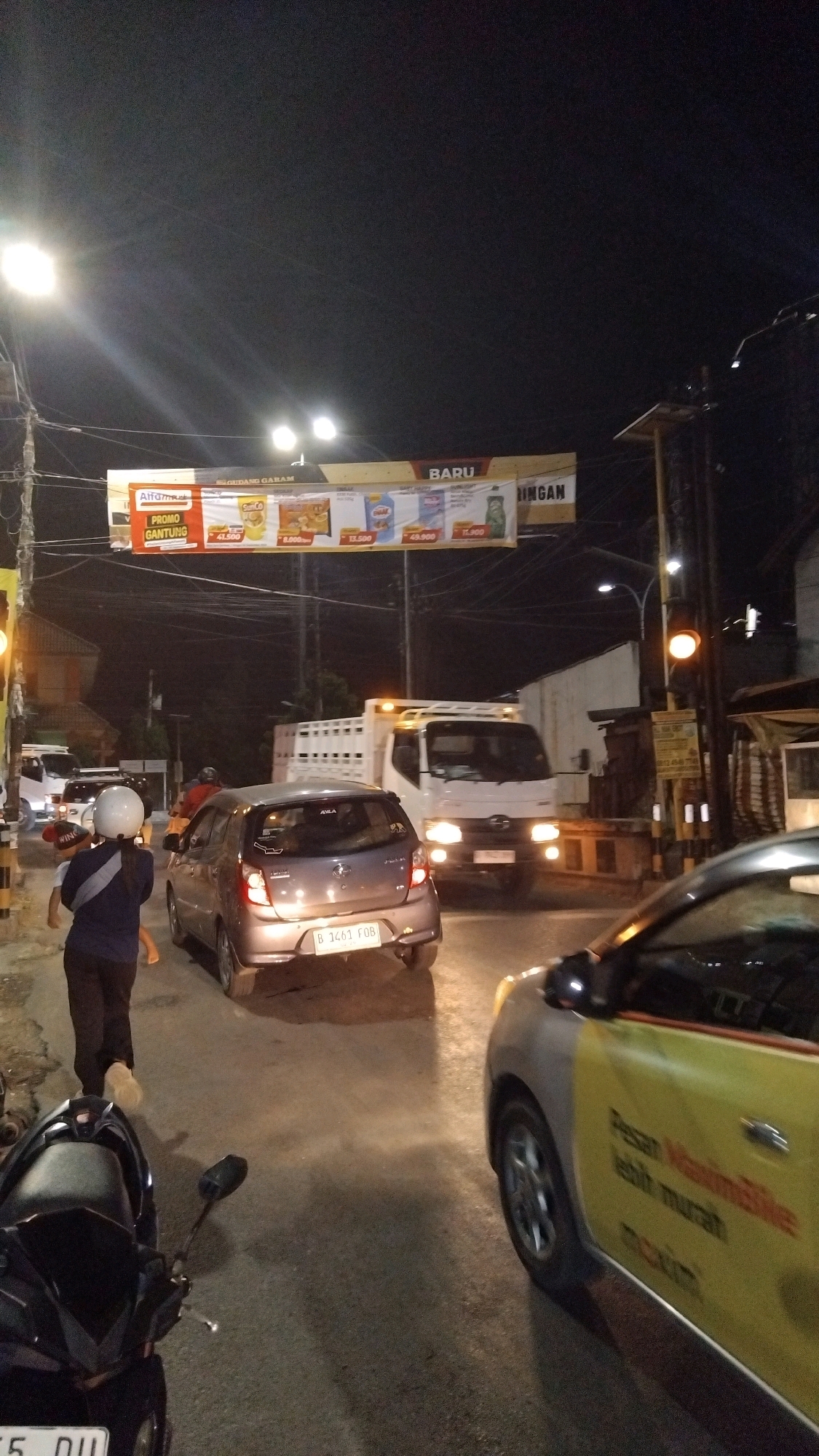 The image depicts a street scene at night in Tegal, Indonesia, likely near a busy intersection. The foreground is dominated by a motorcycle on the left and the side of a yellow and grey car. The car has text on its door that reads "Pesan Maxim Bike lebih murah". In the middle ground, a silver hatchback with license plate "B 1461 FOB" is stopped. A person wearing a dark top, dark pants, and a white helmet is walking away from the viewer towards the left, holding a child's hand. Behind the hatchback, a white truck with an empty cargo bed is stopped. Overhead, a large banner advertises various products, including items from Alfamart and "Gudang Garam" cigarettes, with prices listed below some items. The banner also has the word "BARU" (new) on it. Streetlights and traffic lights illuminate the scene, casting reflections on the wet pavement. The environment appears urban, with buildings and utility poles visible in the background.