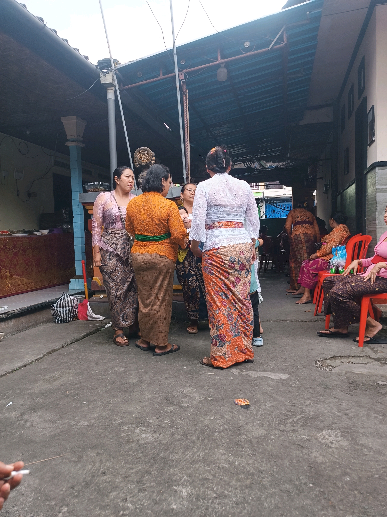 In Tabanan, Indonesia, several women, some wearing traditional Balinese attire like kebaya and sarongs, stand and sit in an outdoor covered area. A few of the women are engaged in conversation, while others are seated on red chairs. One woman in the foreground holds a cigarette. The environment appears to be a street or courtyard with a concrete floor and a corrugated metal roof supported by posts. In the background, there are buildings and some decorative elements, including what appears to be a small shrine.