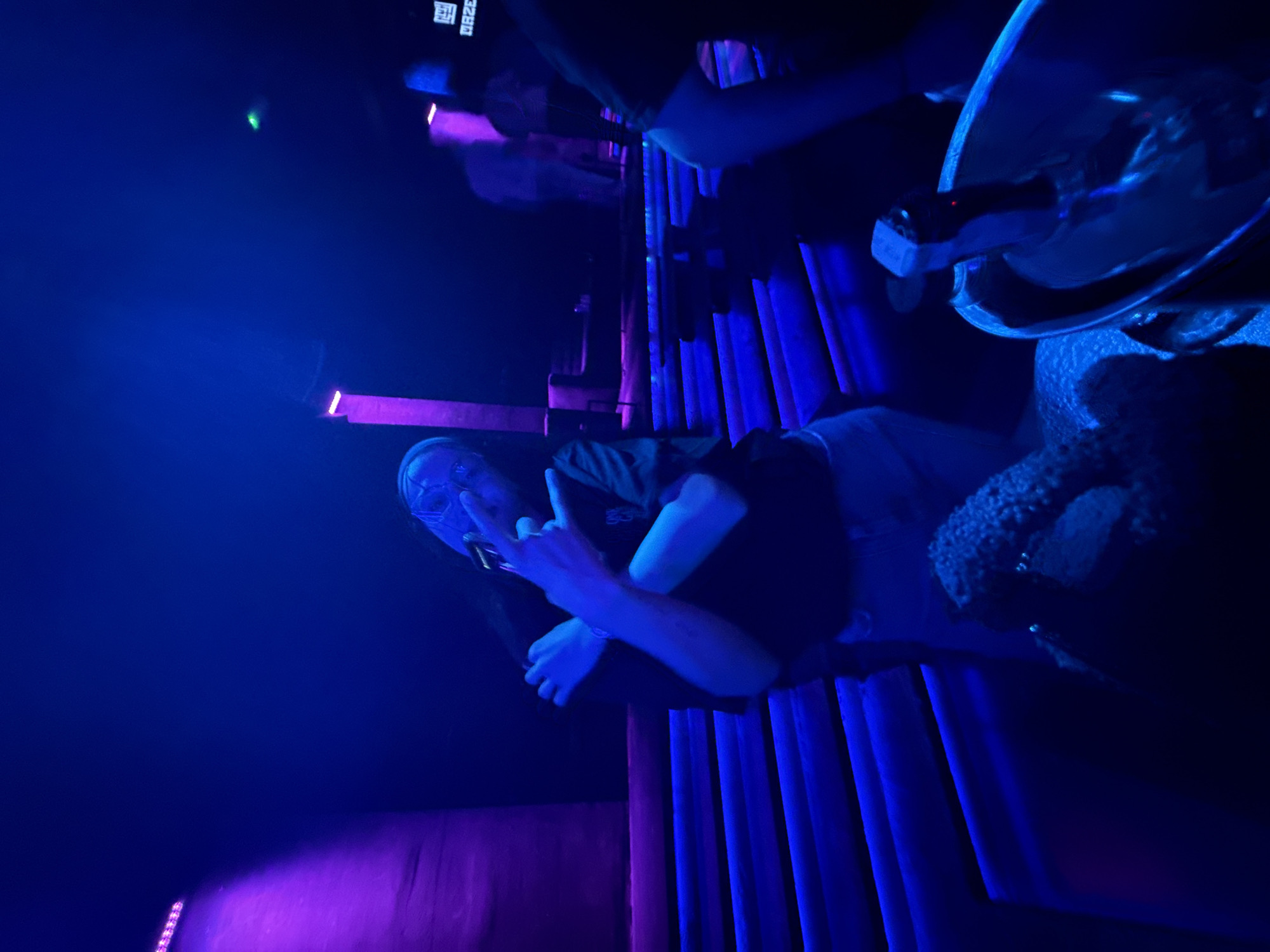 A man wearing a mask holds up two fingers, while standing in front of blue curtains. A bottle of champagne is in an ice bucket, visible in the top right. Purple and blue lighting is in the background. A crowd of people is visible behind the curtains. The location is unknown.